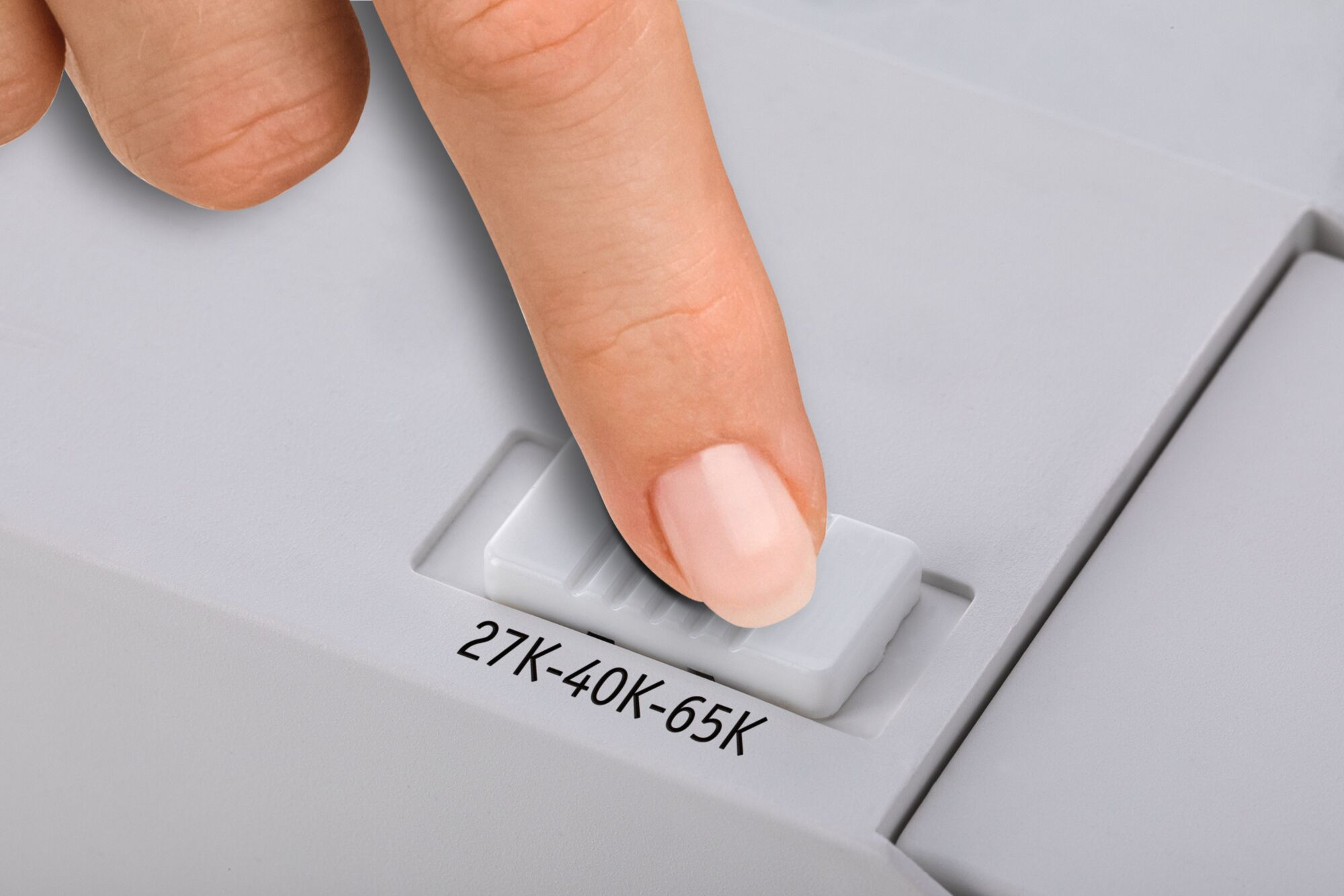 Palec naciska biały suwak z napisem 27K-40K-65K. Suwak znajduje się na jasnoszarej powierzchni.