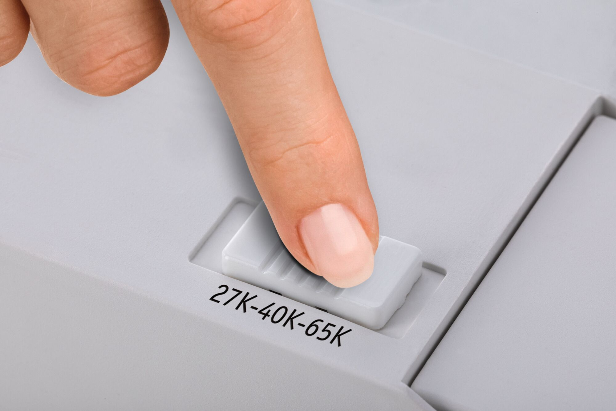 Ein Finger drückt einen weißen Schalter mit der Aufschrift '27K-40K-65K' auf einer hellgrauen Oberfläche.