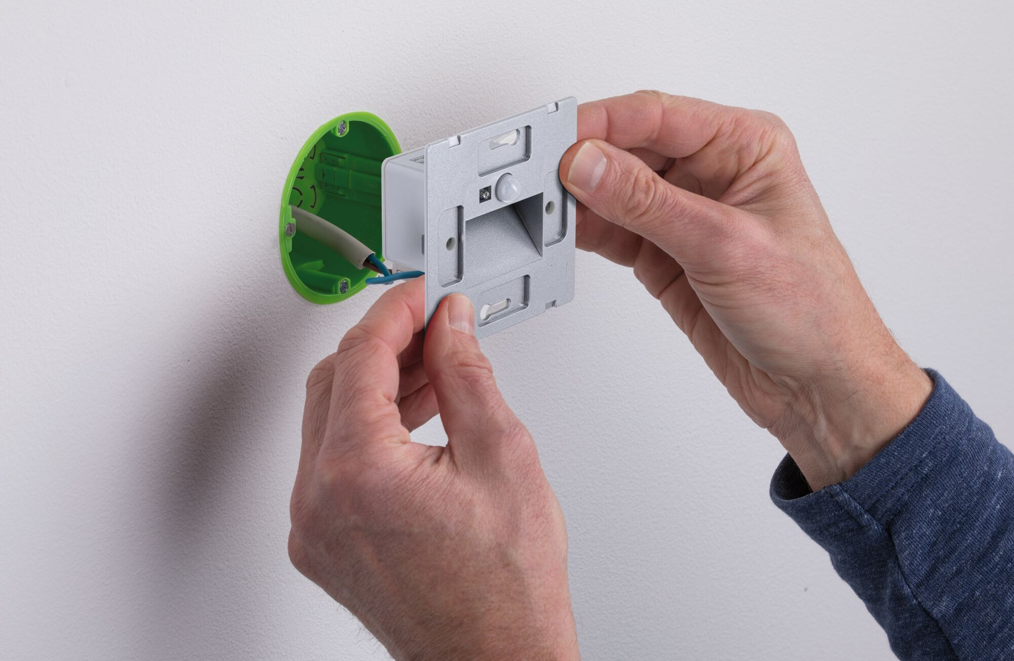 Hände installieren einen Lichtsensor an einer weißen Wand. Eine grüne Elektrobox ist sichtbar. Drähte und ein silberner Sensor sind vorhanden.