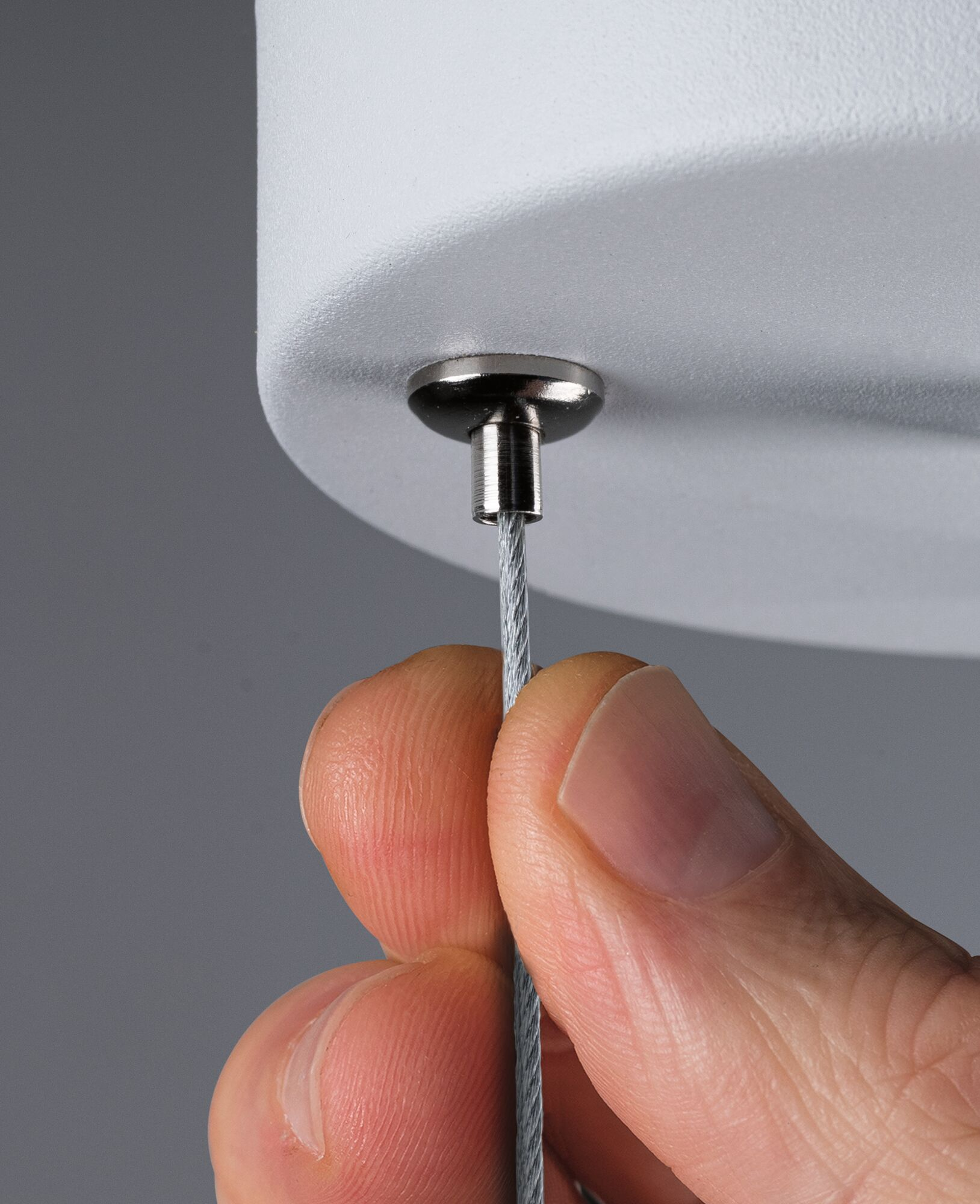 Ręka trzyma stalową linkę przymocowaną do metalowego mocowania. Tło to szara ściana i białe mocowanie.