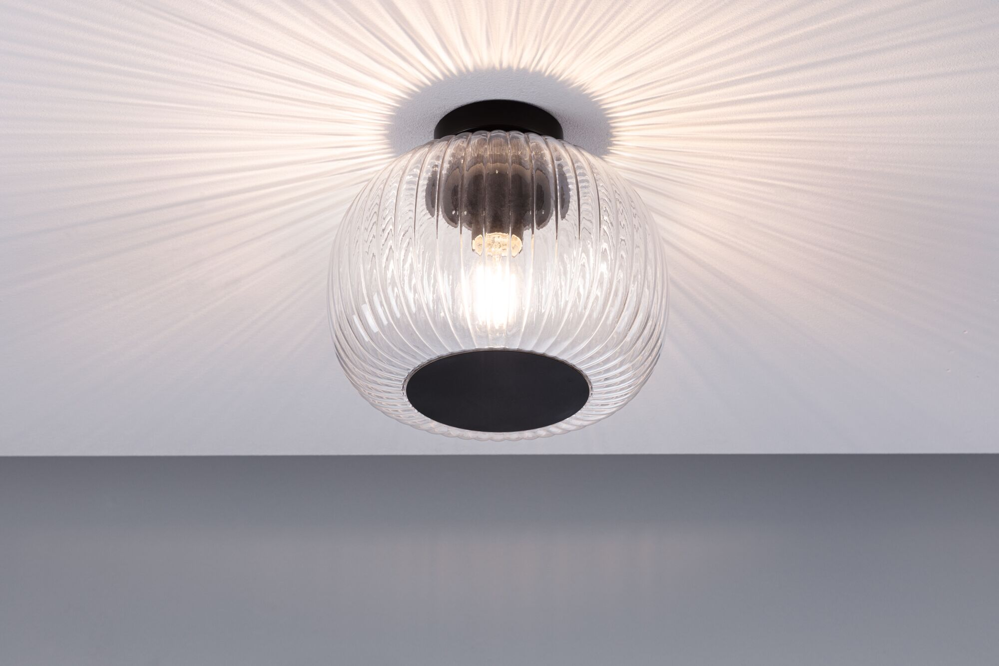 Eine Deckenlampe aus Glas mit schwarzem Sockel hängt an der weißen Decke. Licht wirft Schatten auf die Decke.