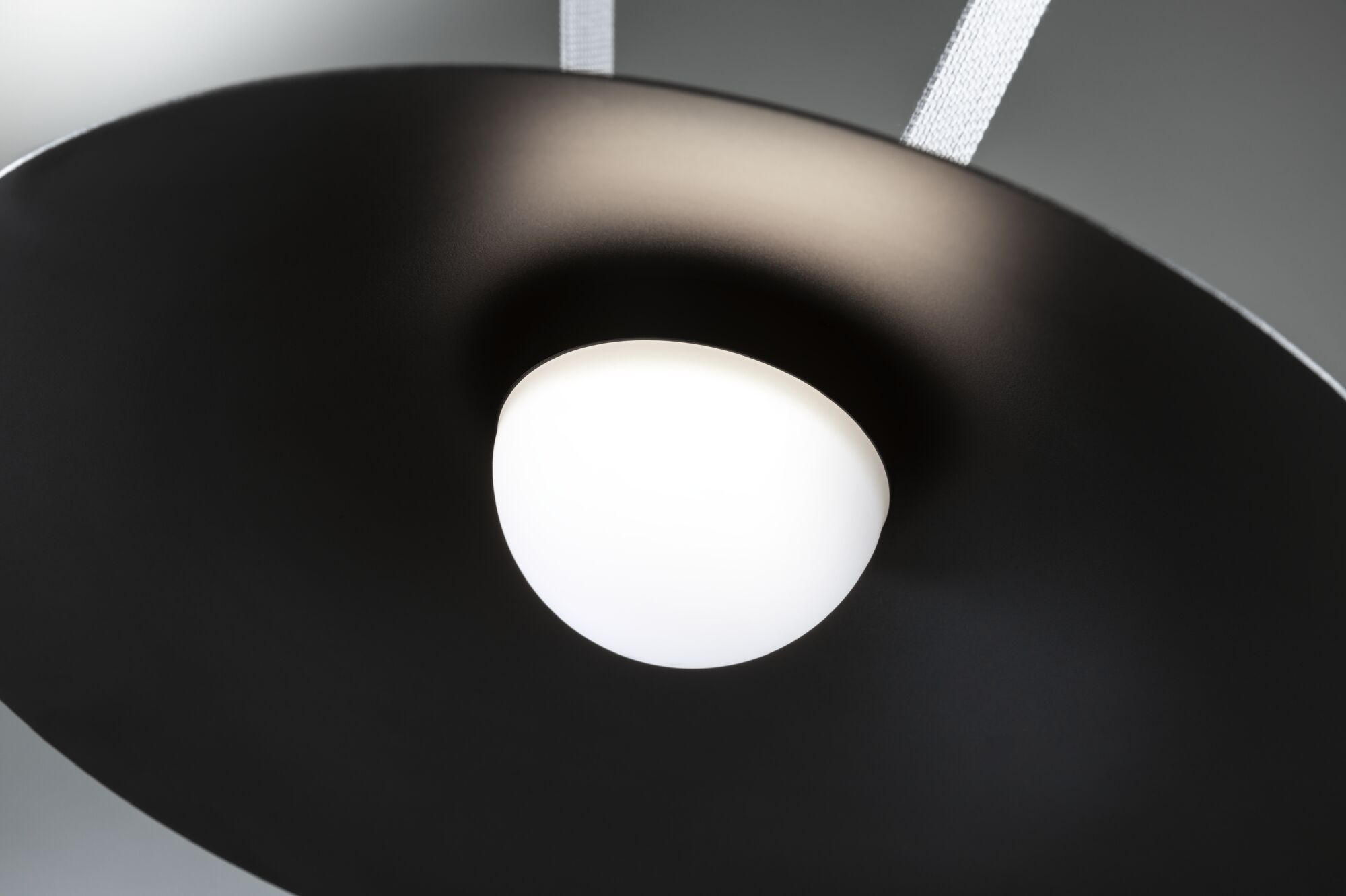 Nowoczesna czarna lampa z białym, świecącym światłem pośrodku, zawieszona na białych paskach materiału, na szarym tle.
