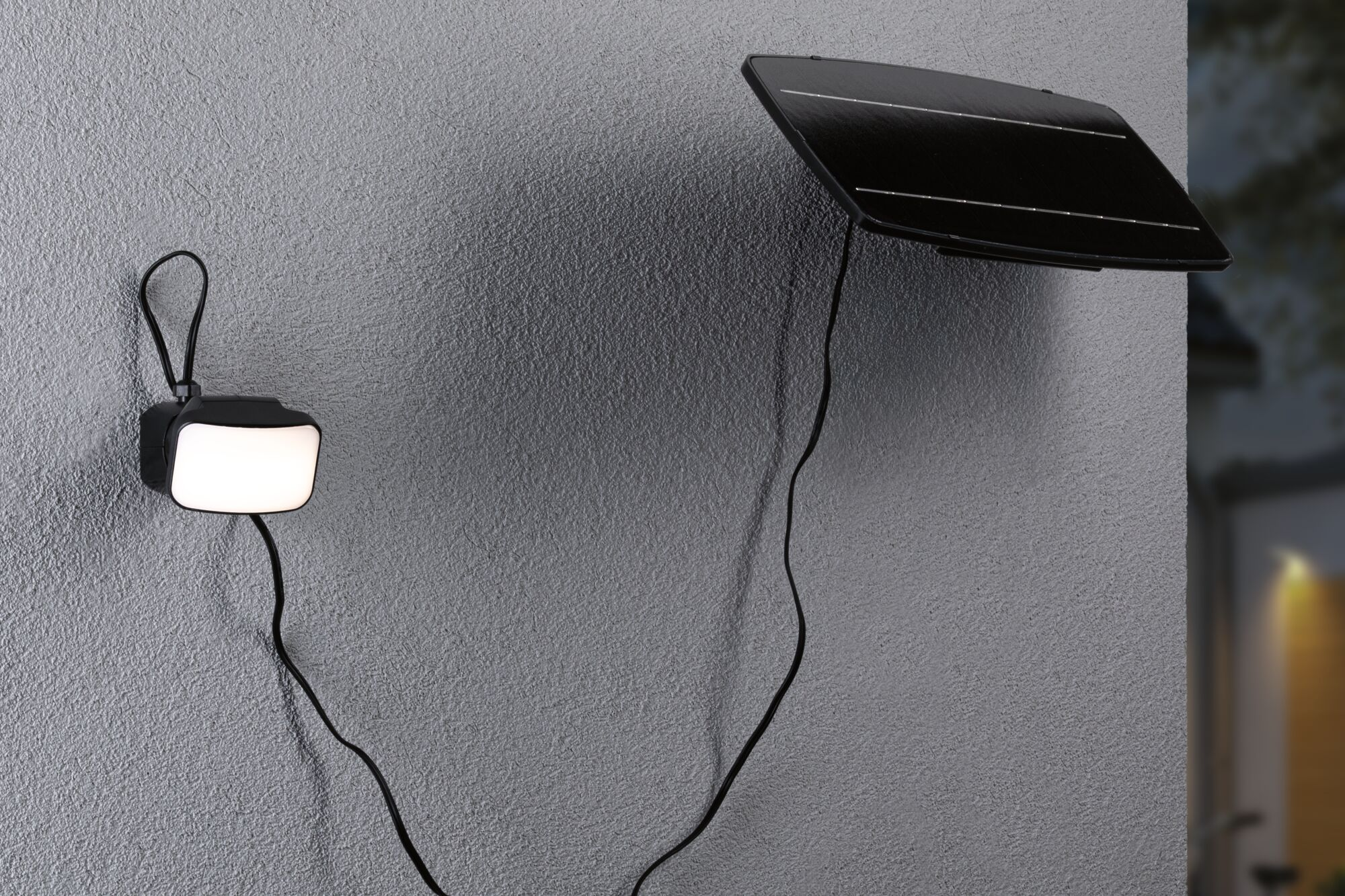 Czarny panel słoneczny i kwadratowa lampa zamontowane na szarej, teksturowanej ścianie. Kable je łączą.
