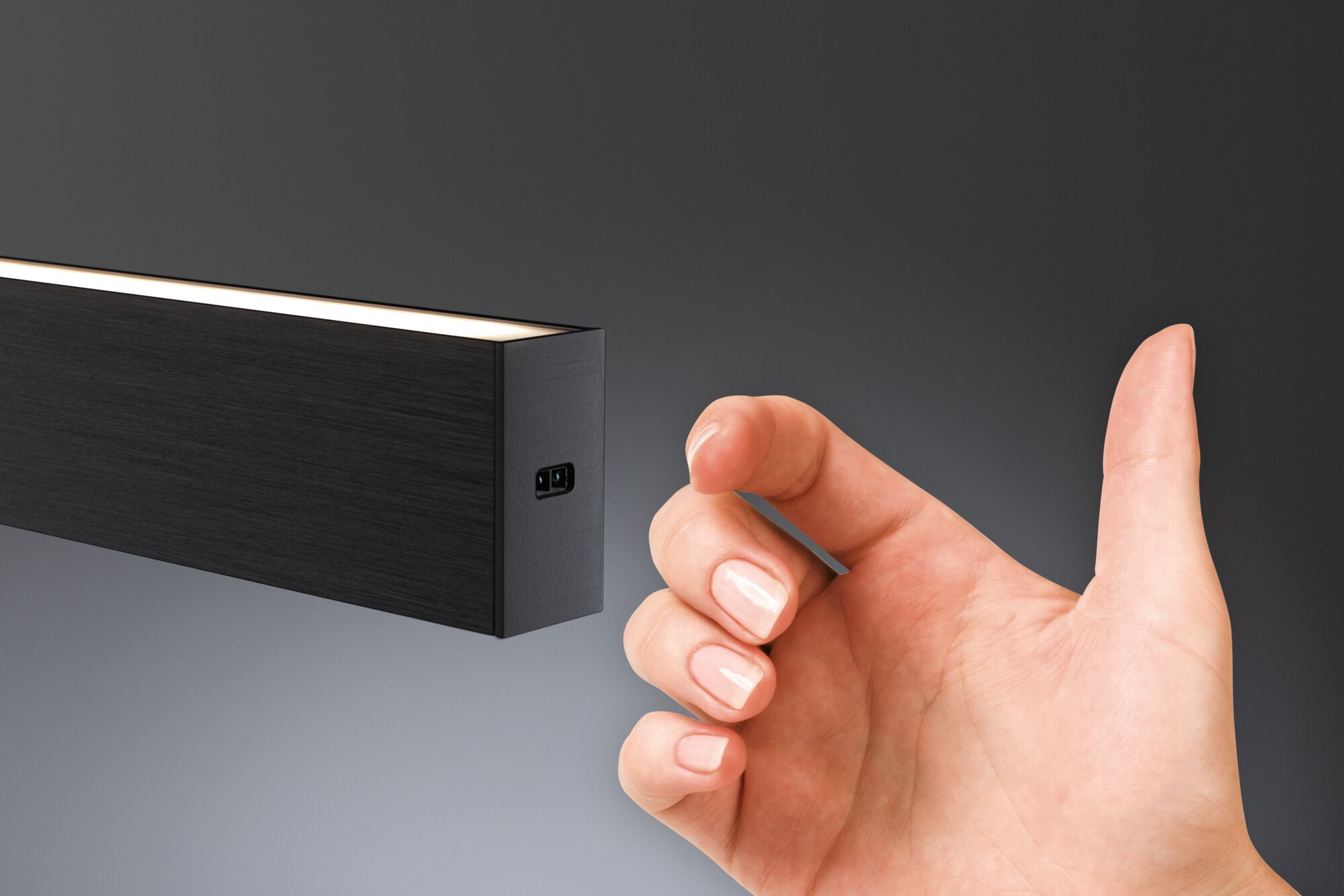 Dłoń gestykuluje w stronę ciemnej, prostokątnej oprawy oświetleniowej z podświetlanym wnętrzem, na szarym tle gradientowym.