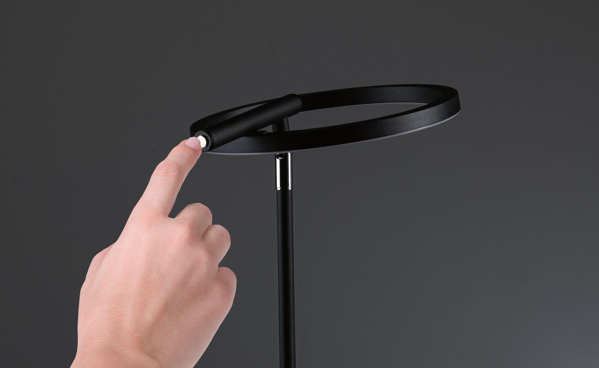 Dłoń dotyka czarnej lampy pierścieniowej, palcem naciskając przycisk zasilania. Tło jest szare.
