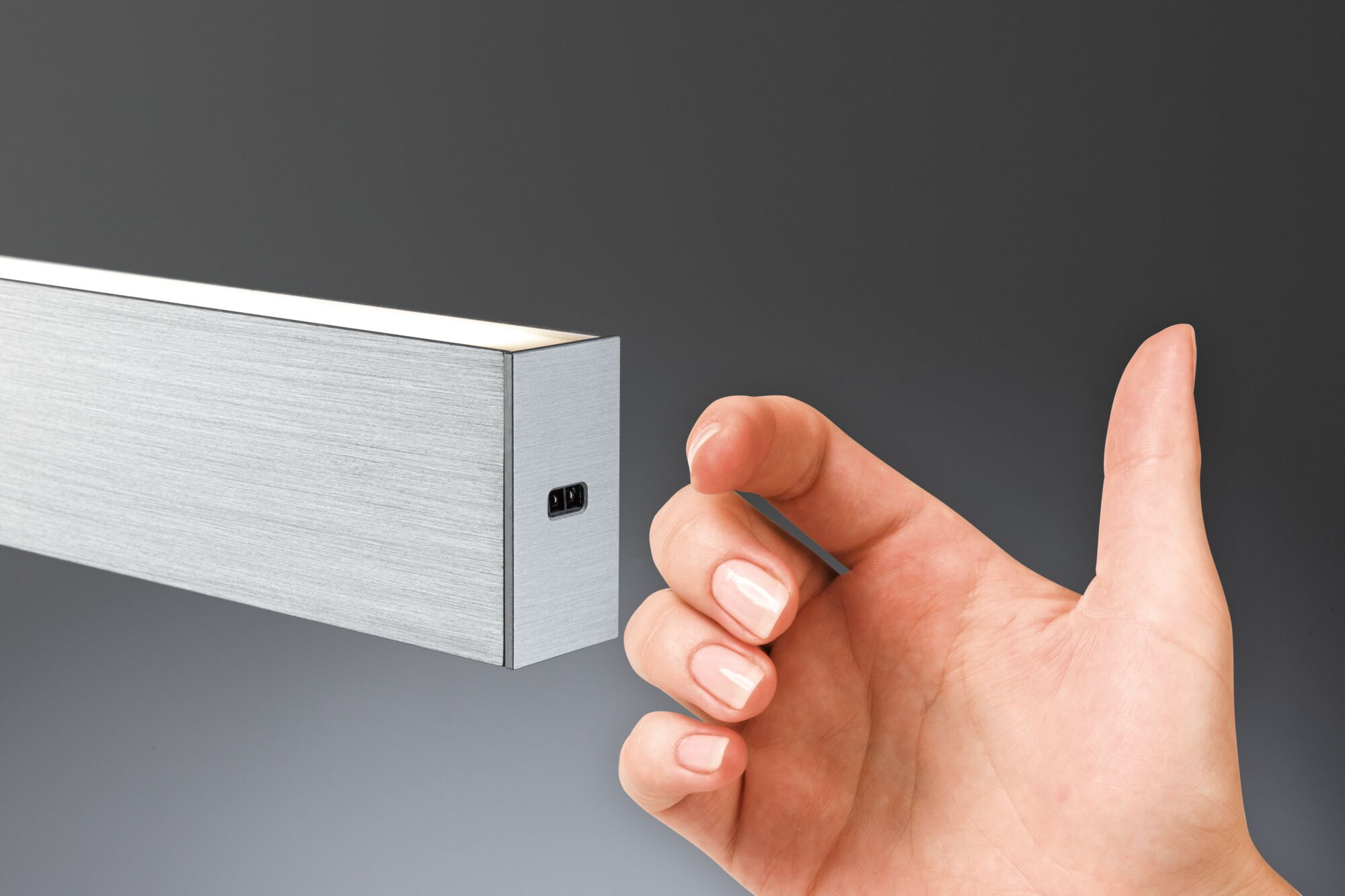 Eine Hand mit gepflegten Nägeln zeigt auf eine metallische Leuchte. Der Hintergrund ist ein Grauton.