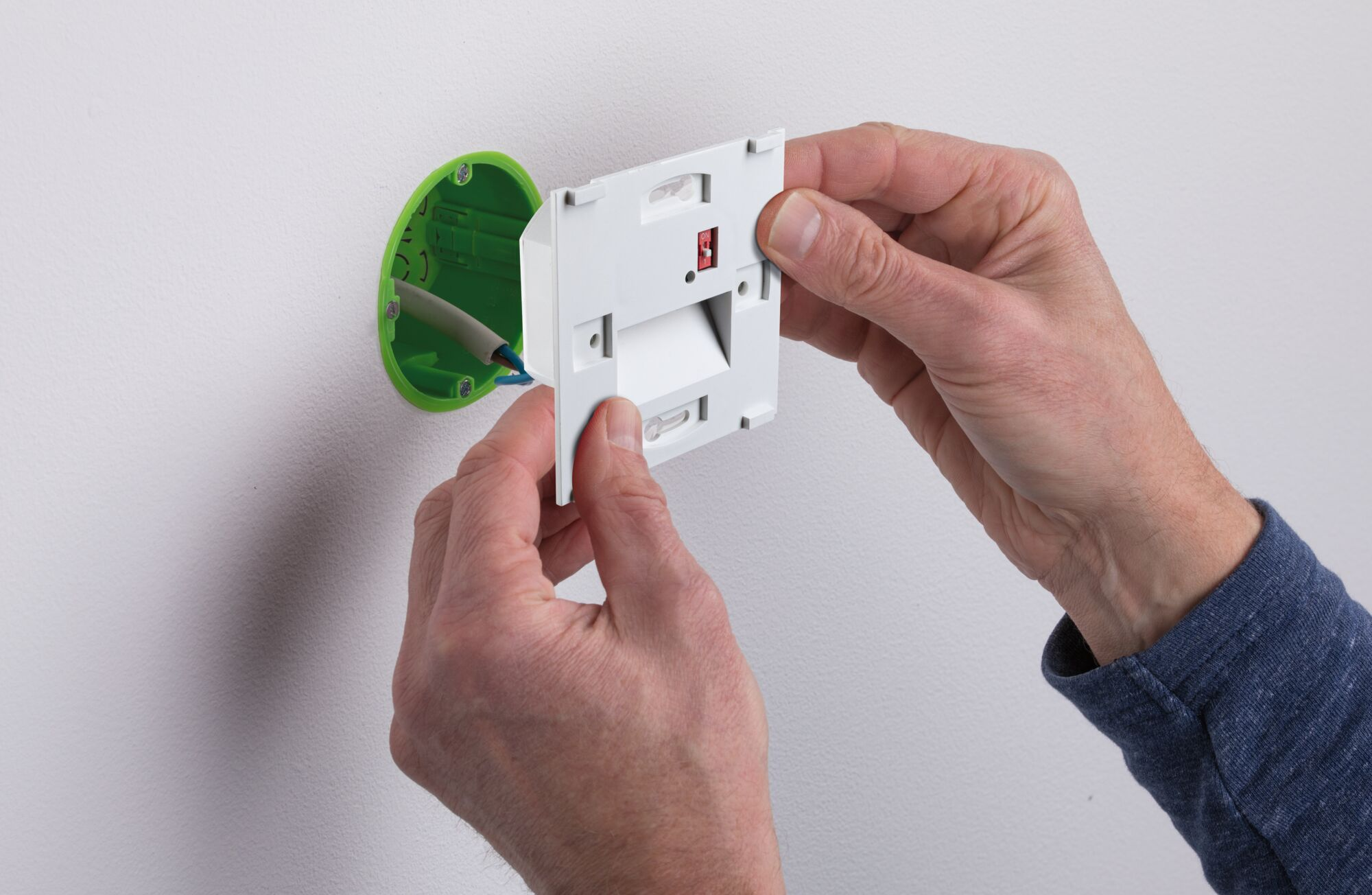 Hände installieren eine weiße elektrische Komponente an einer Wand, neben einer grünen Einbaubox mit Drähten.