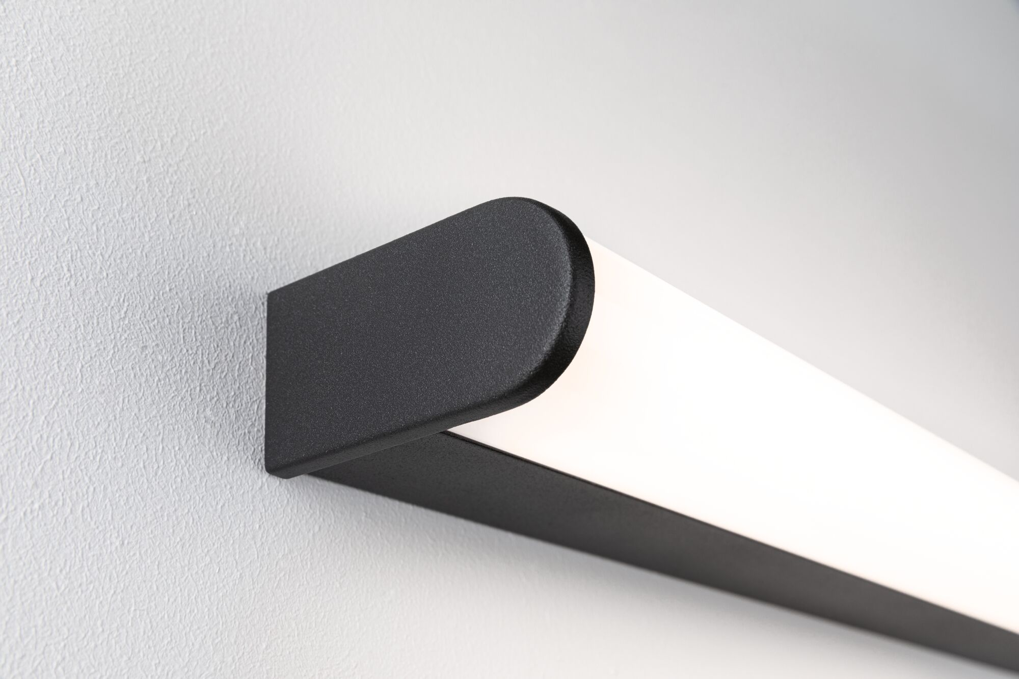 Eine schwarze, rechteckige Leuchte ist an einer strukturierten, hellgrauen Wand angebracht. Das Licht emittiert ein helles, weißes Leuchten.