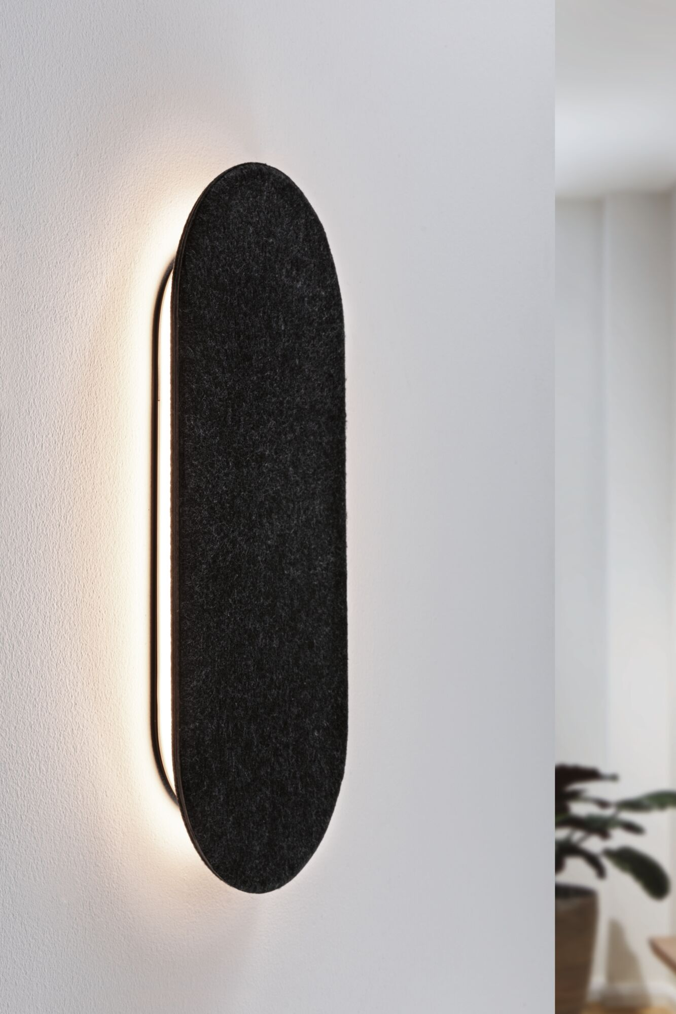 Czarna, owalna lampa ścienna o fakturze filcu, zamontowana na białej ścianie. Światło emanuje z boku.