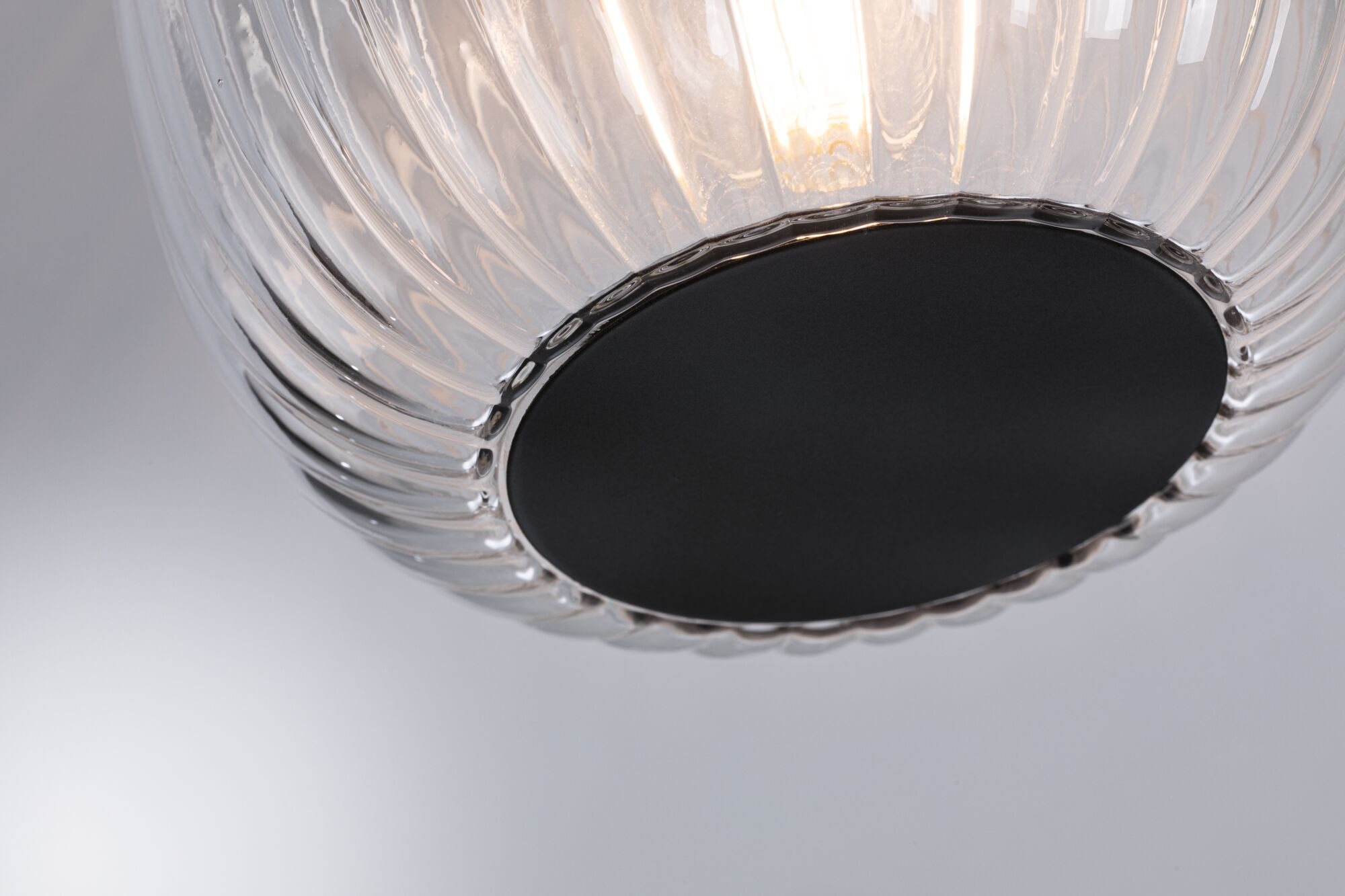 Nahaufnahme einer klaren Glaslampe mit schwarzem Sockel. Geripptes Glasdesign. Helles Licht scheint durch die Mitte der Lampe.