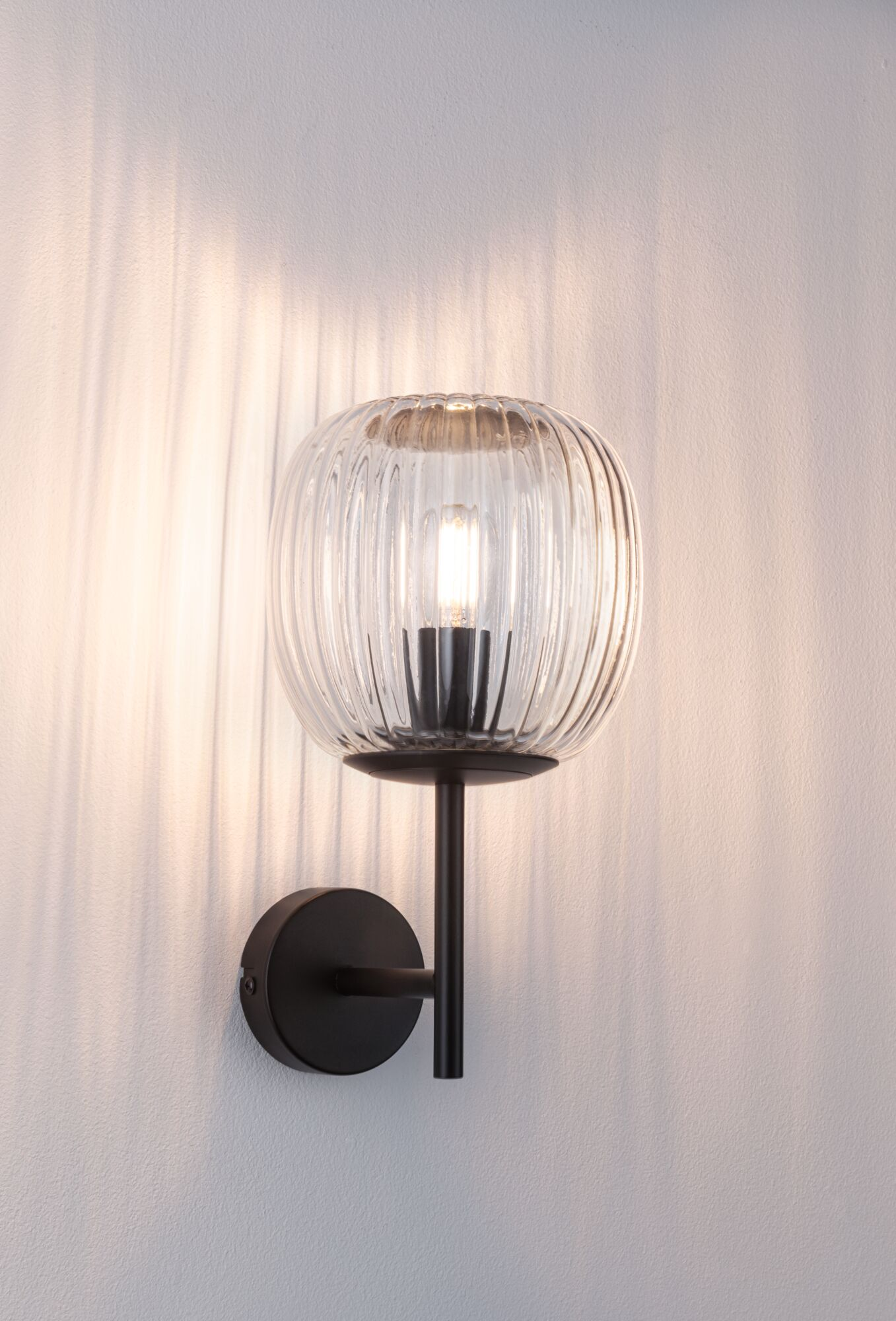 Eine Wandlampe mit einem transparenten, gerippten Glasschirm und einem schwarzen Metallsockel. Das Licht wirft Schatten auf die weiße Wand.