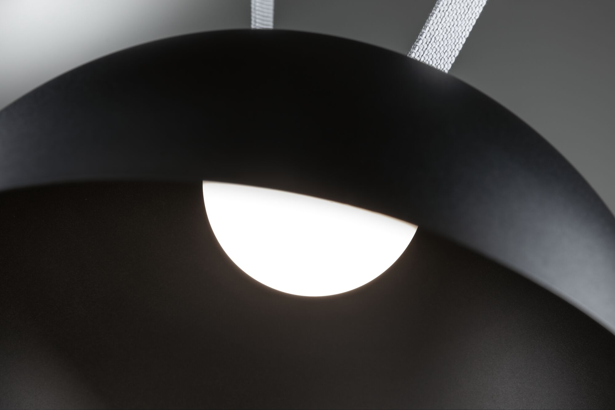 Eine schwarze, gebogene Lampe mit einer hellen, weißen Lichtquelle. Weißer Riemen oben. Grauer Hintergrund.