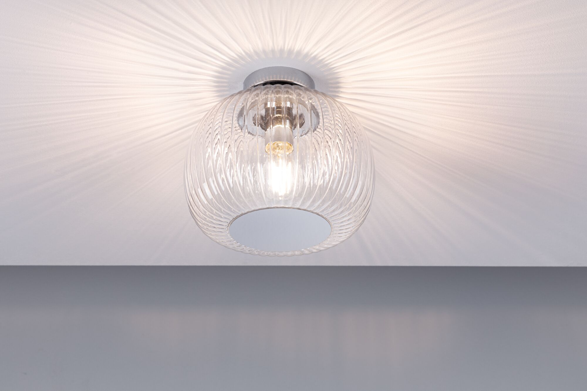 Lampa sufitowa z przezroczystym, wzorzystym kloszem. Światło promieniuje z niego, tworząc cienie na białym suficie.