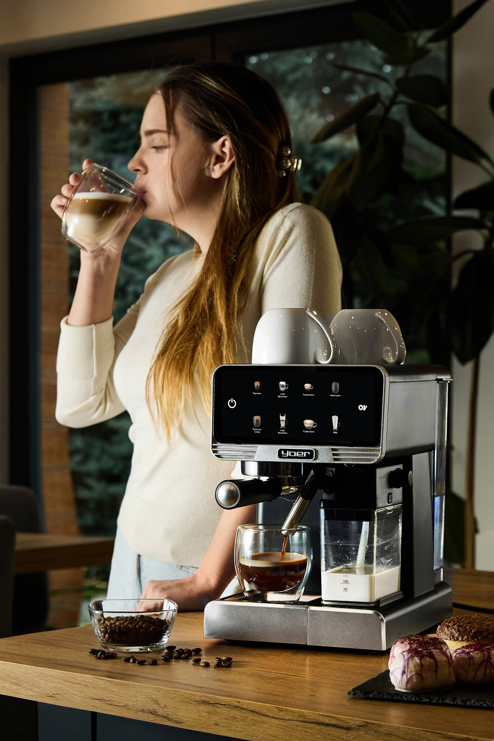 Kobieta pije kawę ze szklanki. Na stole stoi ekspres do kawy. Obecne są również ziarna kawy i pączki.