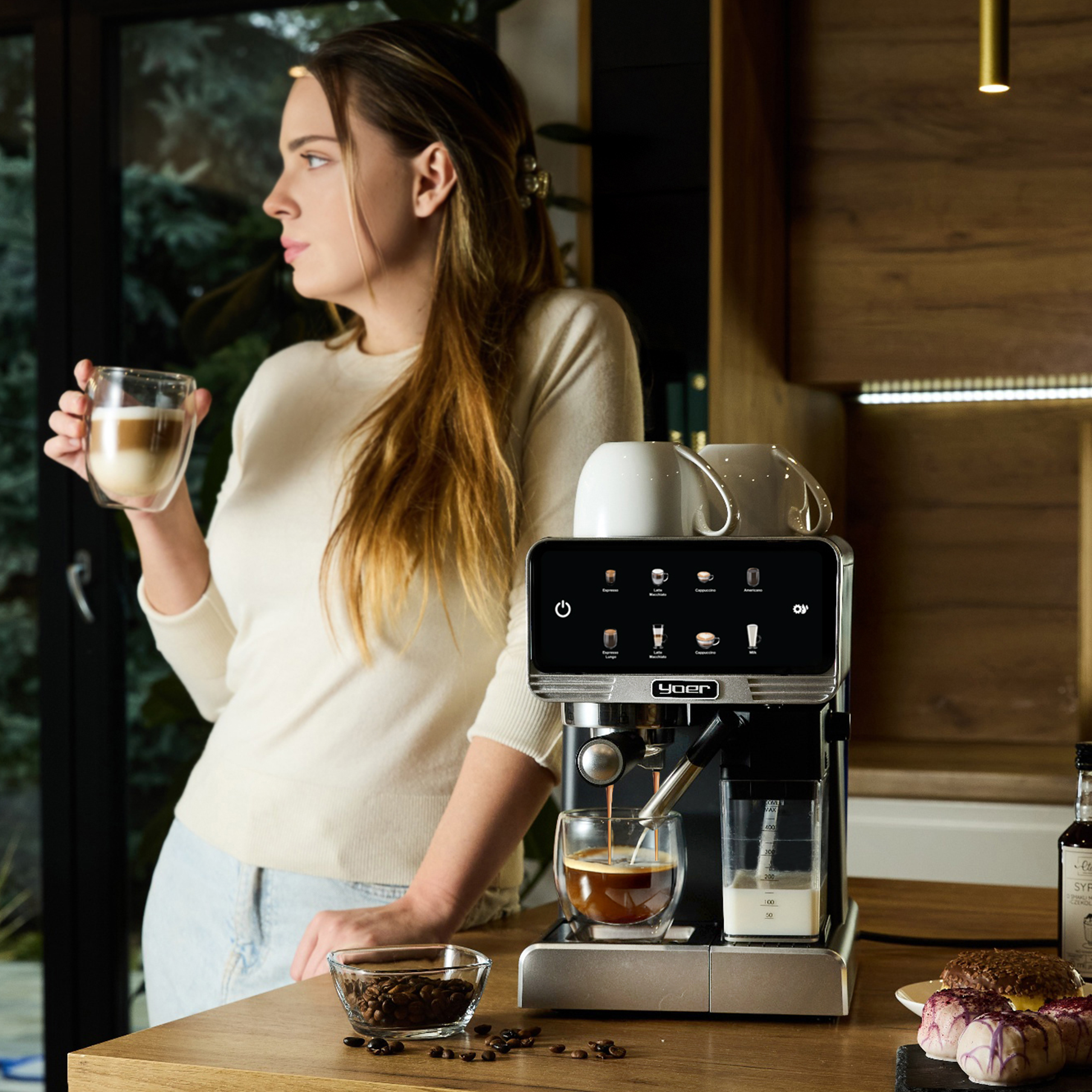 Kobieta trzymająca filiżankę kawy, spogląda przez okno. Ekspres do kawy i ciastka na blacie.