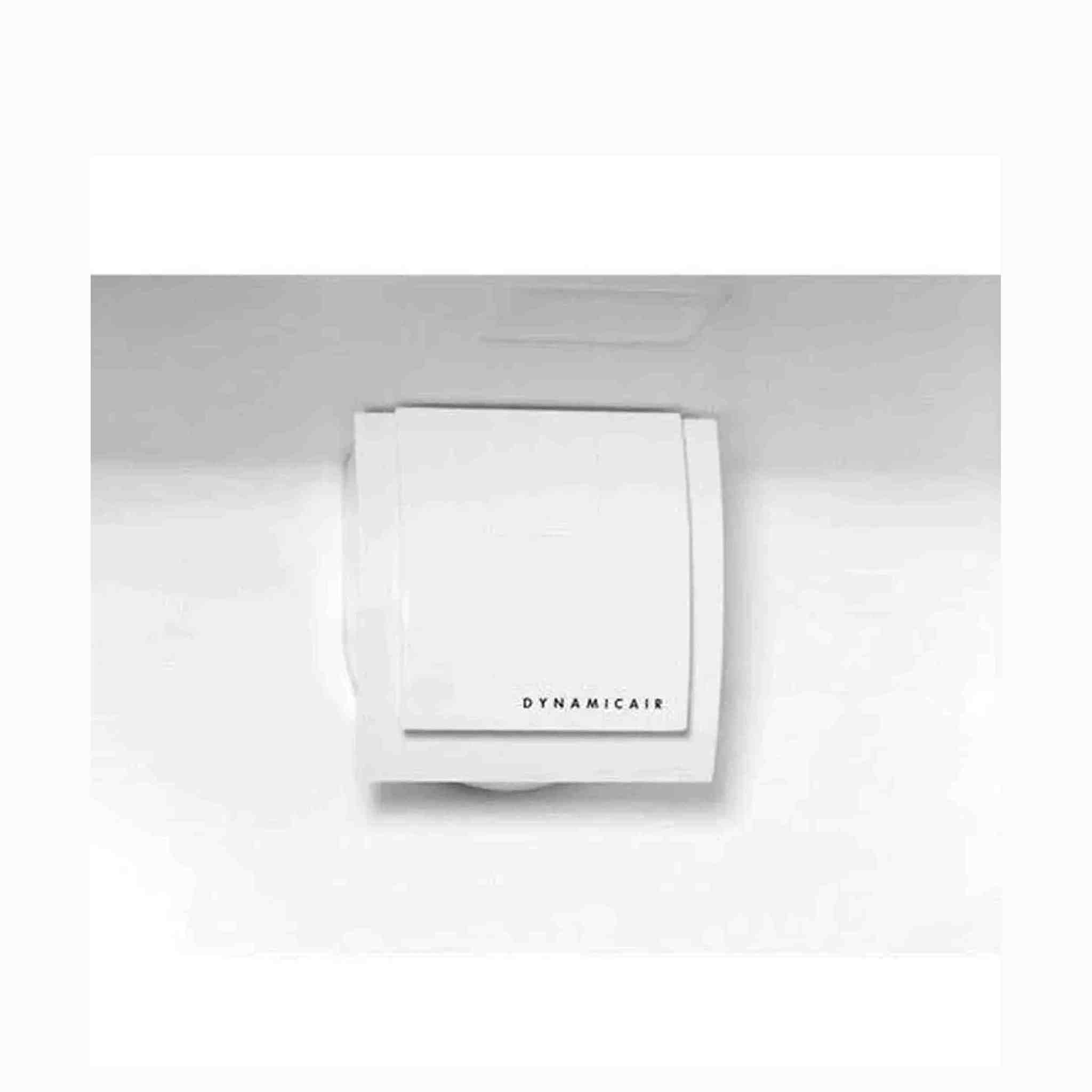 A white Dynamicair air vent is mounted on a white surface. The device has a square shape with a central panel.