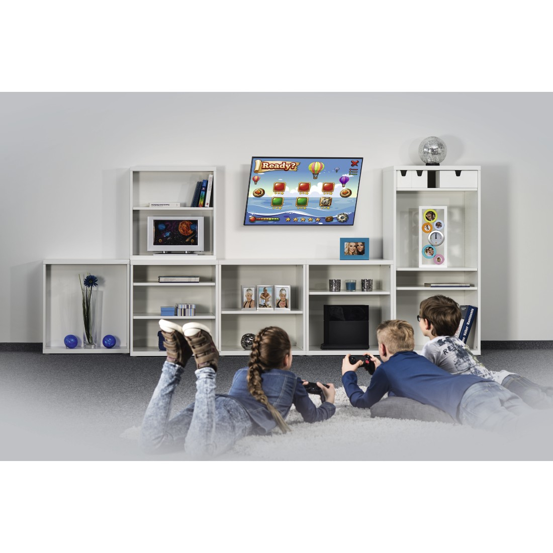 Tres niños jugando videojuegos en el suelo. Un televisor muestra un juego. Estanterías blancas y decoración en el fondo.