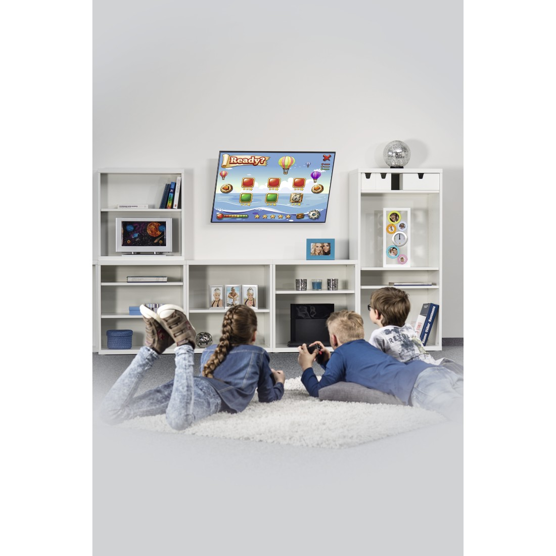 Troje dzieci leży na dywanie i gra w grę wideo na telewizorze. Telewizor wyświetla kolorową grę. Otaczają je półki z książkami i dekoracje.