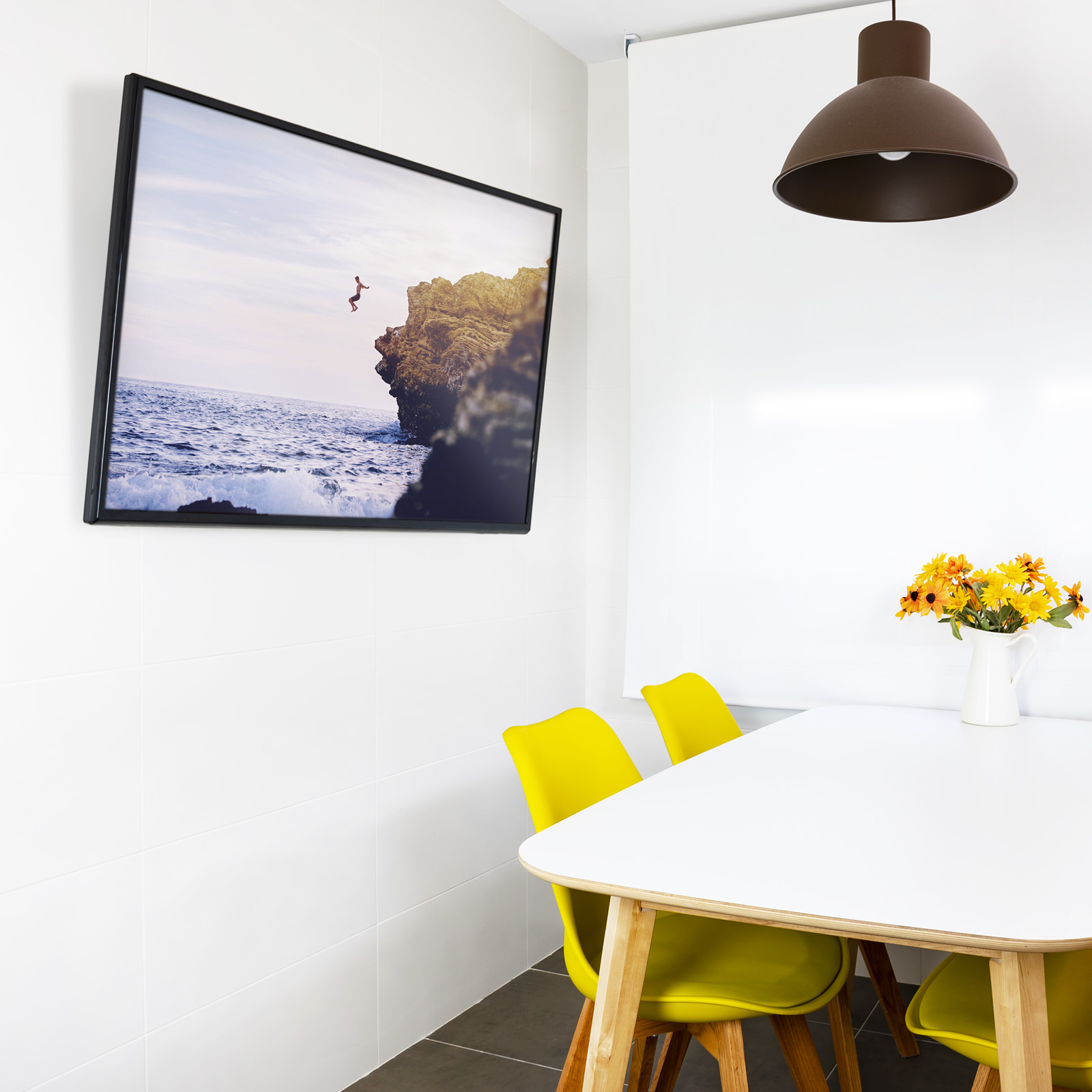 Habitación con foto de persona saltando al mar, mesa blanca, sillas amarillas y lámpara marrón.