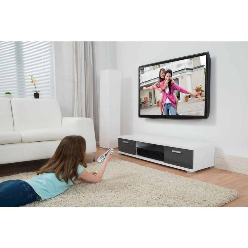 Ein Mädchen liegt auf einem Teppich und sieht fern. Im Fernsehen sind zwei Frauen zu sehen. Ein weißes Sofa und ein Schrank sind im Raum.