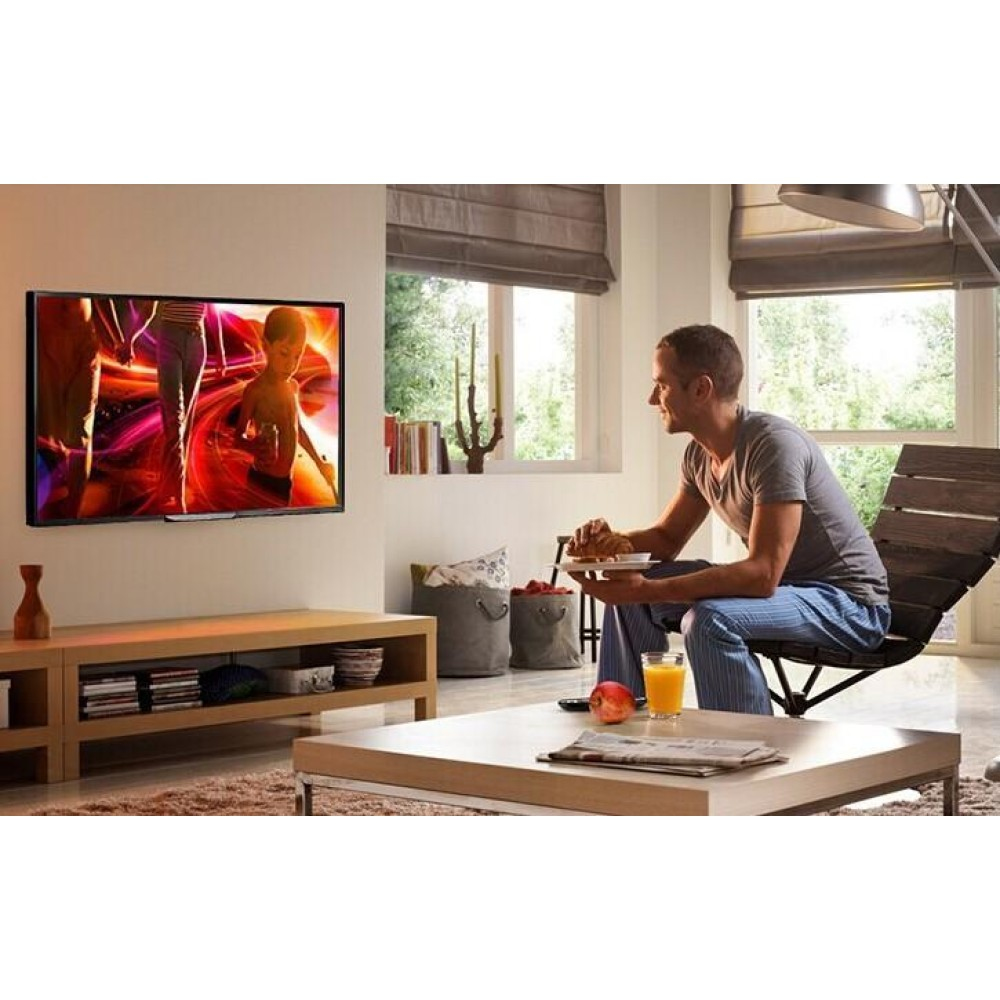 Ein Mann sitzt und isst. Ein Fernseher zeigt ein farbenfrohes Bild. Es gibt einen Tisch mit Saft und Obst. Ein Fenster und ein Holzstuhl sind sichtbar.