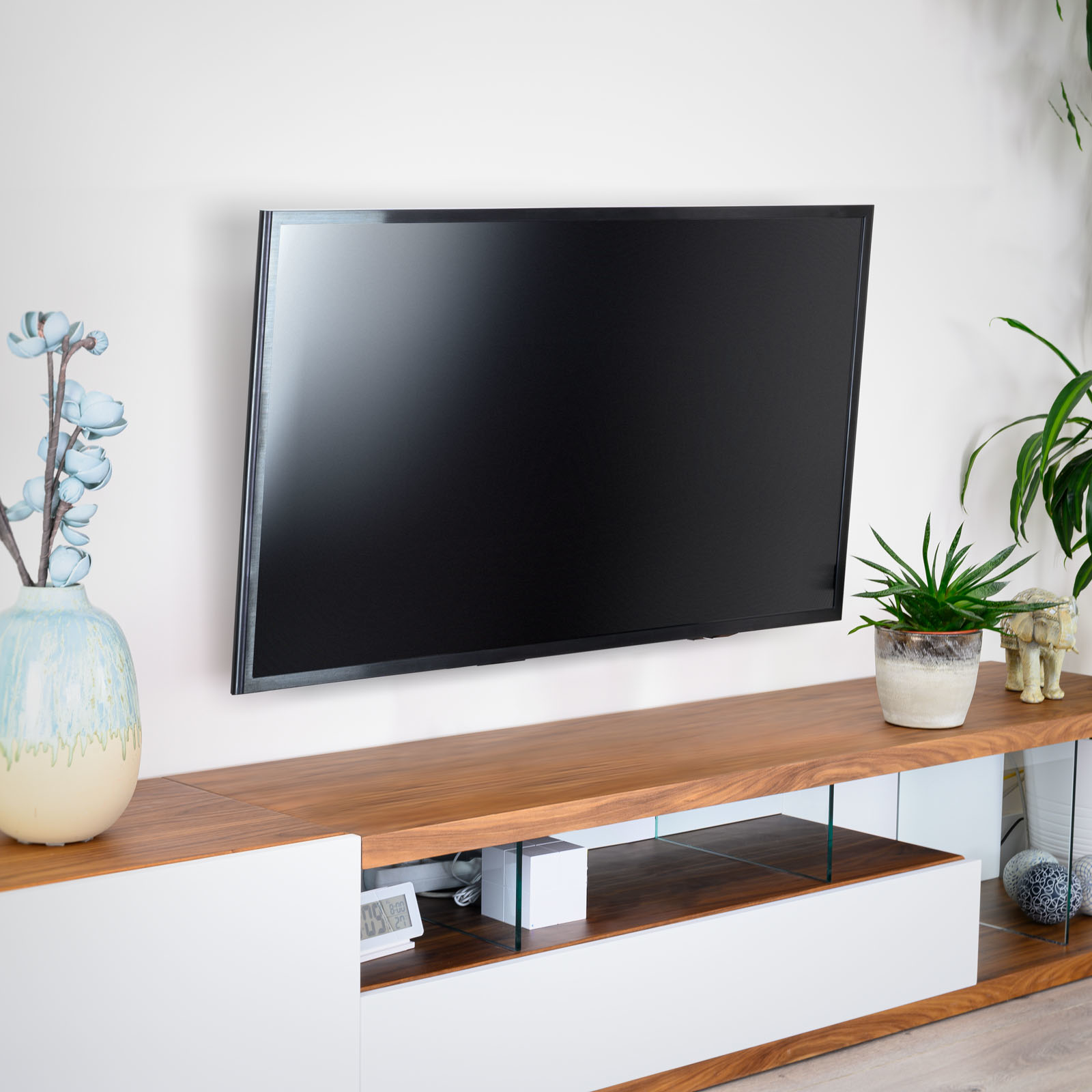 Un televisor montado en una pared blanca sobre un mueble de madera con plantas y decoración. Un jarrón con flores azules a la izquierda.