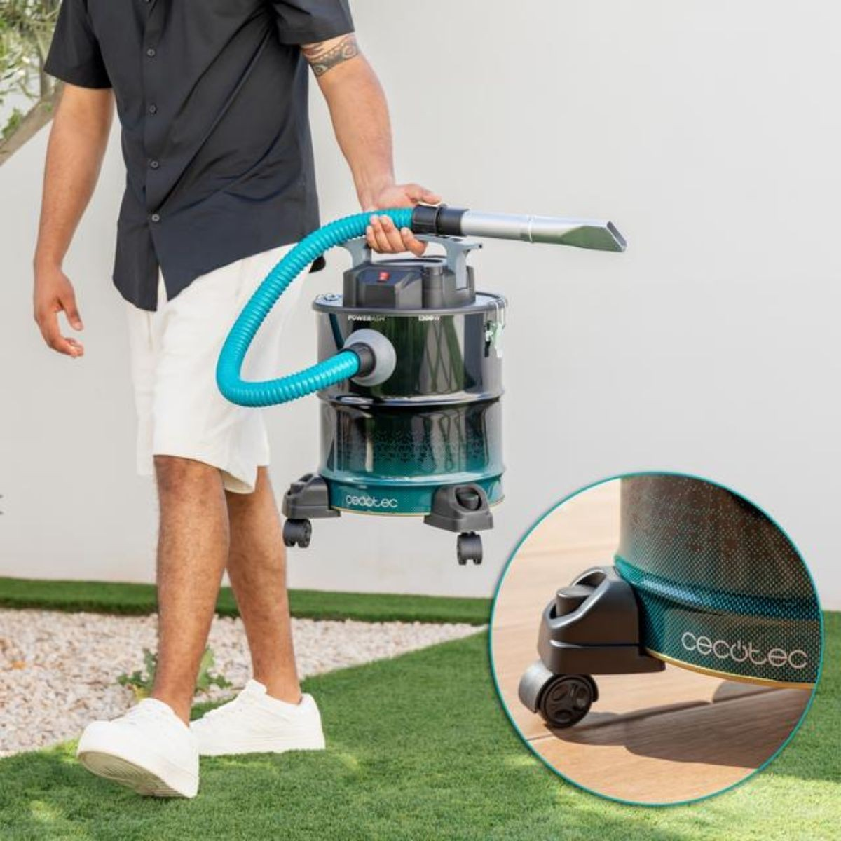 Un uomo tiene un aspirapolvere con un tubo blu. Indossa una camicia nera, pantaloncini bianchi e scarpe da ginnastica. L'aspirapolvere ha le ruote.