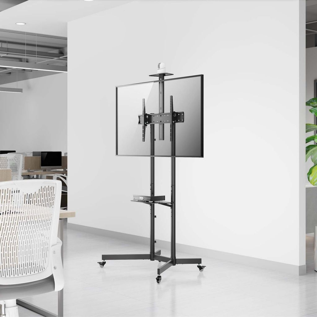 Un soporte de TV negro con una pantalla grande está en una oficina. El soporte tiene ruedas. Una silla y un escritorio blancos están en el fondo.