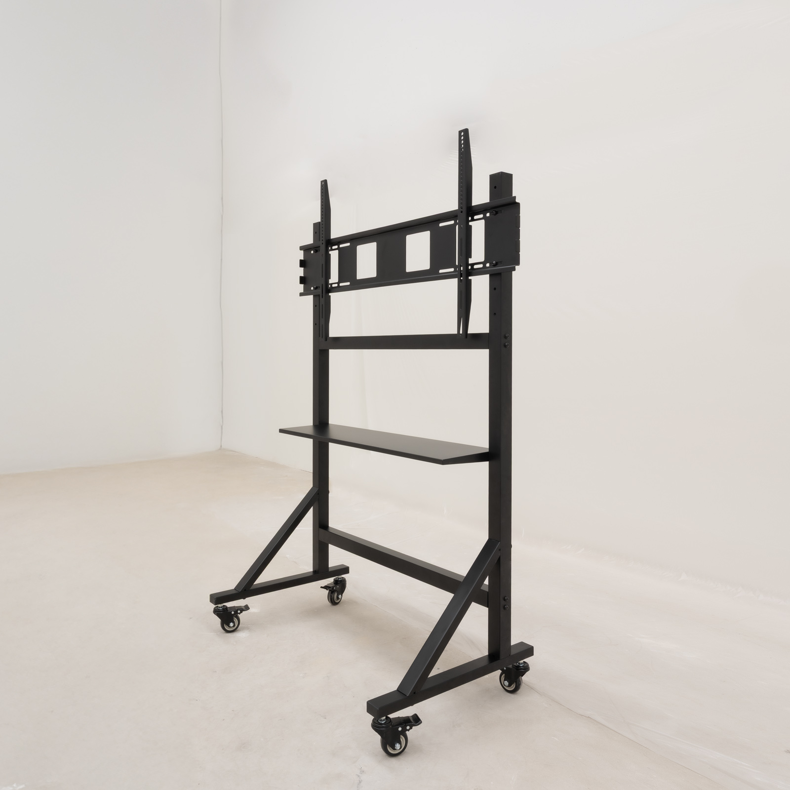 Un soporte de TV de metal negro sobre ruedas, con un estante y soportes de montaje, está en una habitación blanca. El fondo es una pared blanca.
