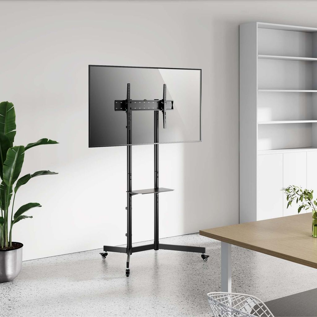 Un televisor negro grande sobre un soporte rodante. Una planta y una mesa son visibles. Paredes blancas y un armario blanco al fondo.