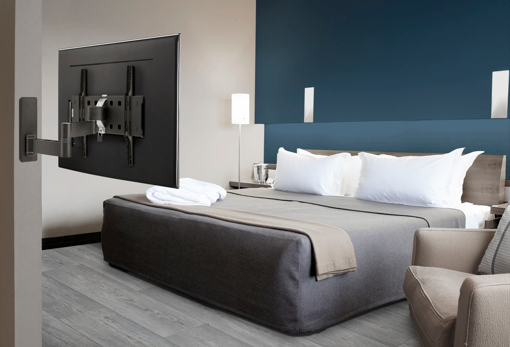 Une chambre d'hôtel avec un lit, une télévision et une chaise. Le lit a des oreillers blancs et une couverture grise. La télévision est fixée au mur.
