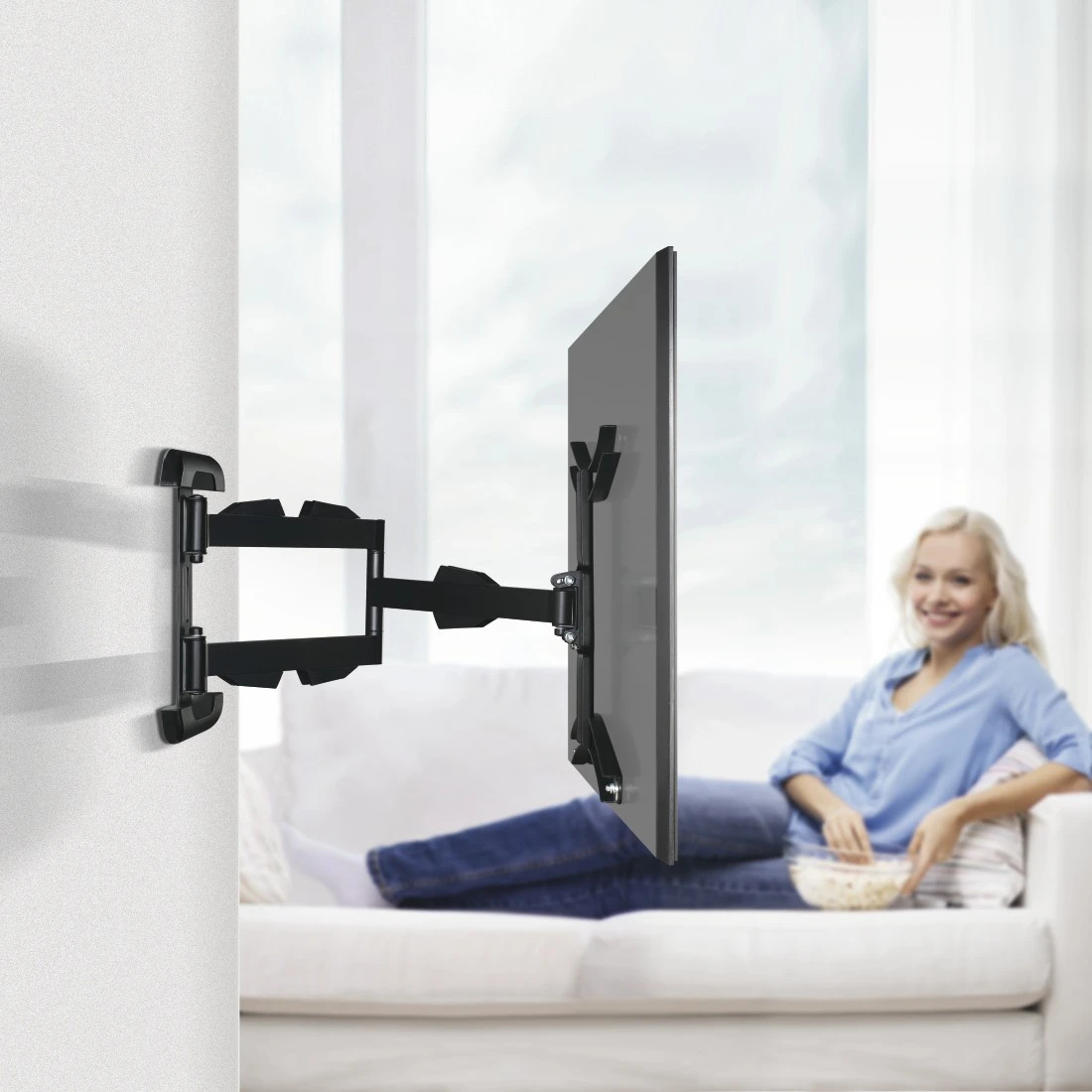 Ein Fernseher mit schwarzer Halterung an der Wand. Eine Frau auf dem Sofa hält eine Schüssel Popcorn.