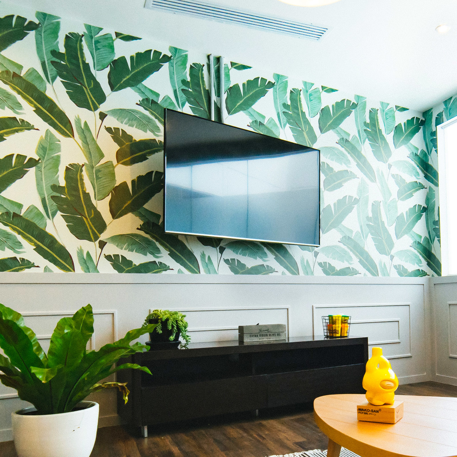 Una habitación con un televisor en una pared con papel pintado de hojas. Un armario negro y una mesa redonda. Se ven plantas en macetas.