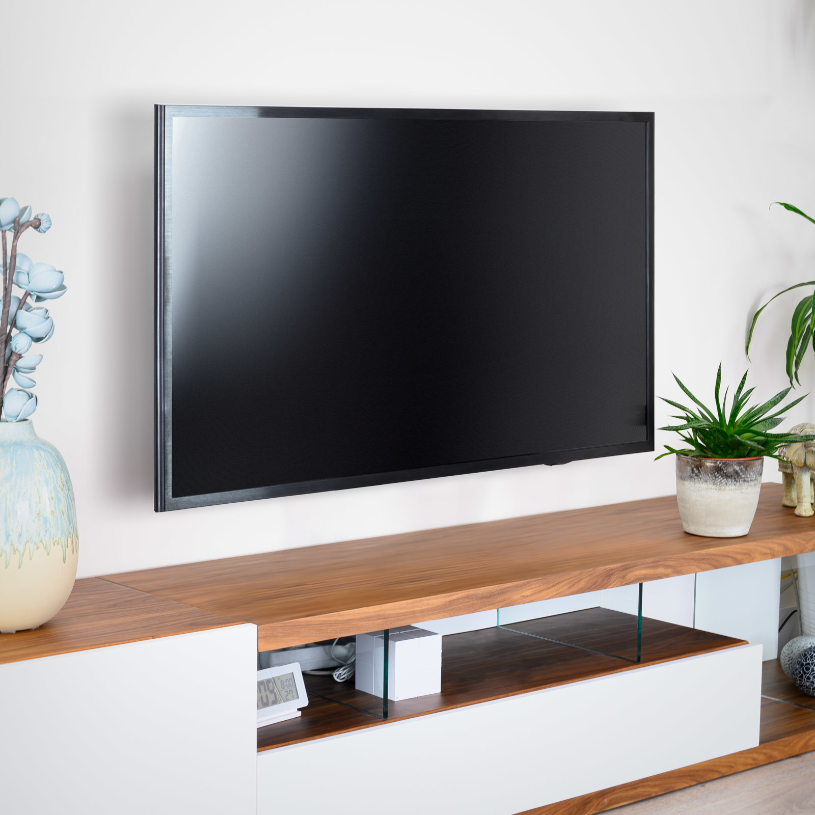 Un televisor negro cuelga sobre un mueble de madera con plantas y decoraciones. Una pared blanca es el fondo.