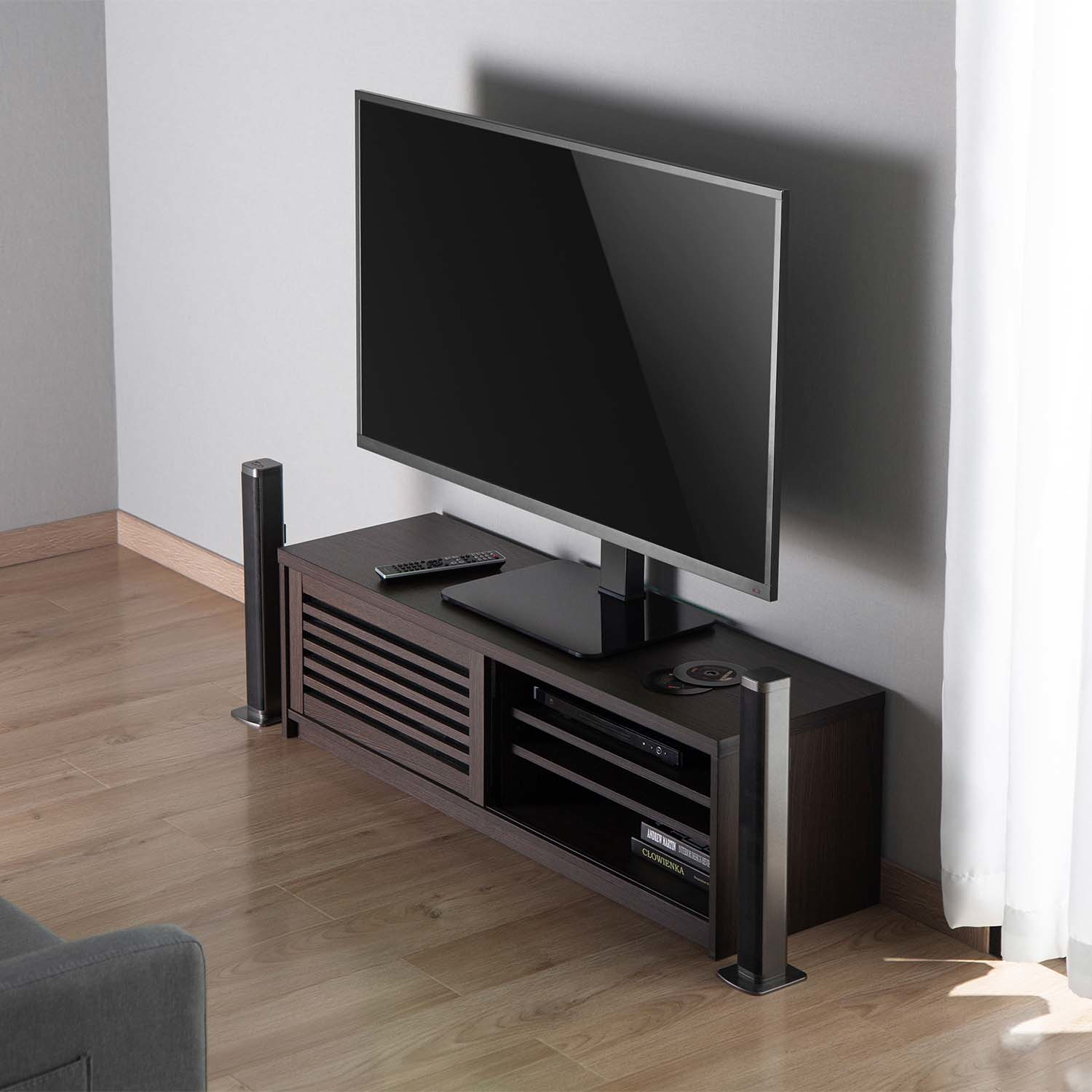 Un televisor de pantalla plana sobre un soporte de madera oscura. Altavoces a ambos lados. Suelo de madera. Pared gris claro. Mando a distancia arriba.