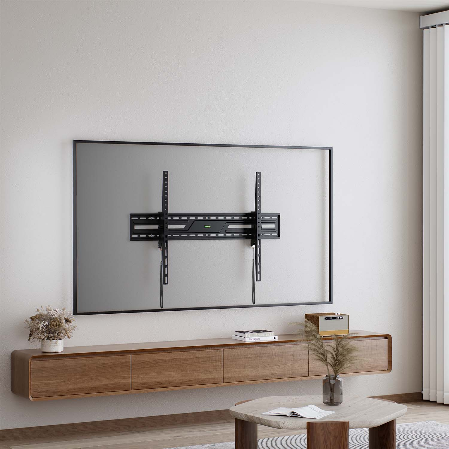 Un televisor montado en la pared con marco negro, un mueble de madera, una mesa de centro y objetos decorativos. Pared blanca y cortinas en el fondo.