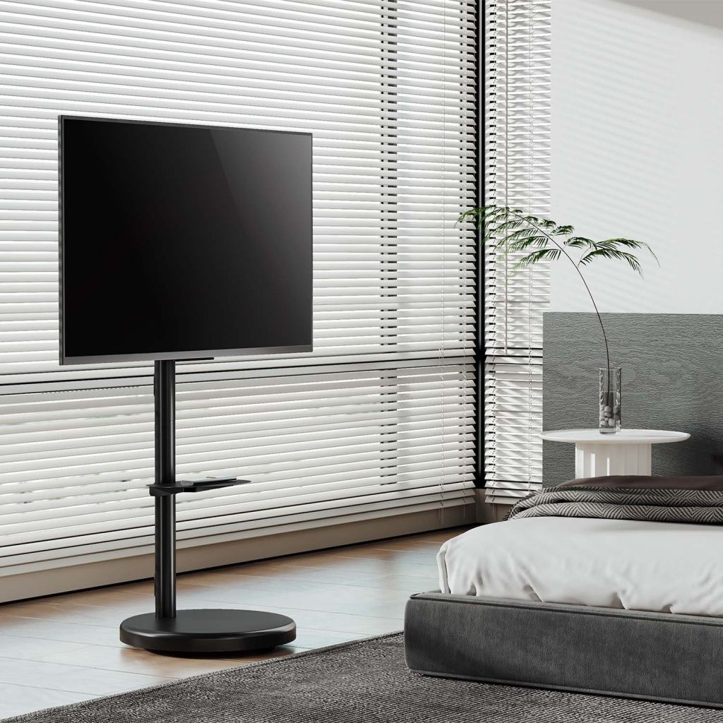 Un televisor negro grande sobre un soporte negro. Persianas, una cama y una mesita con una planta también están en la habitación.