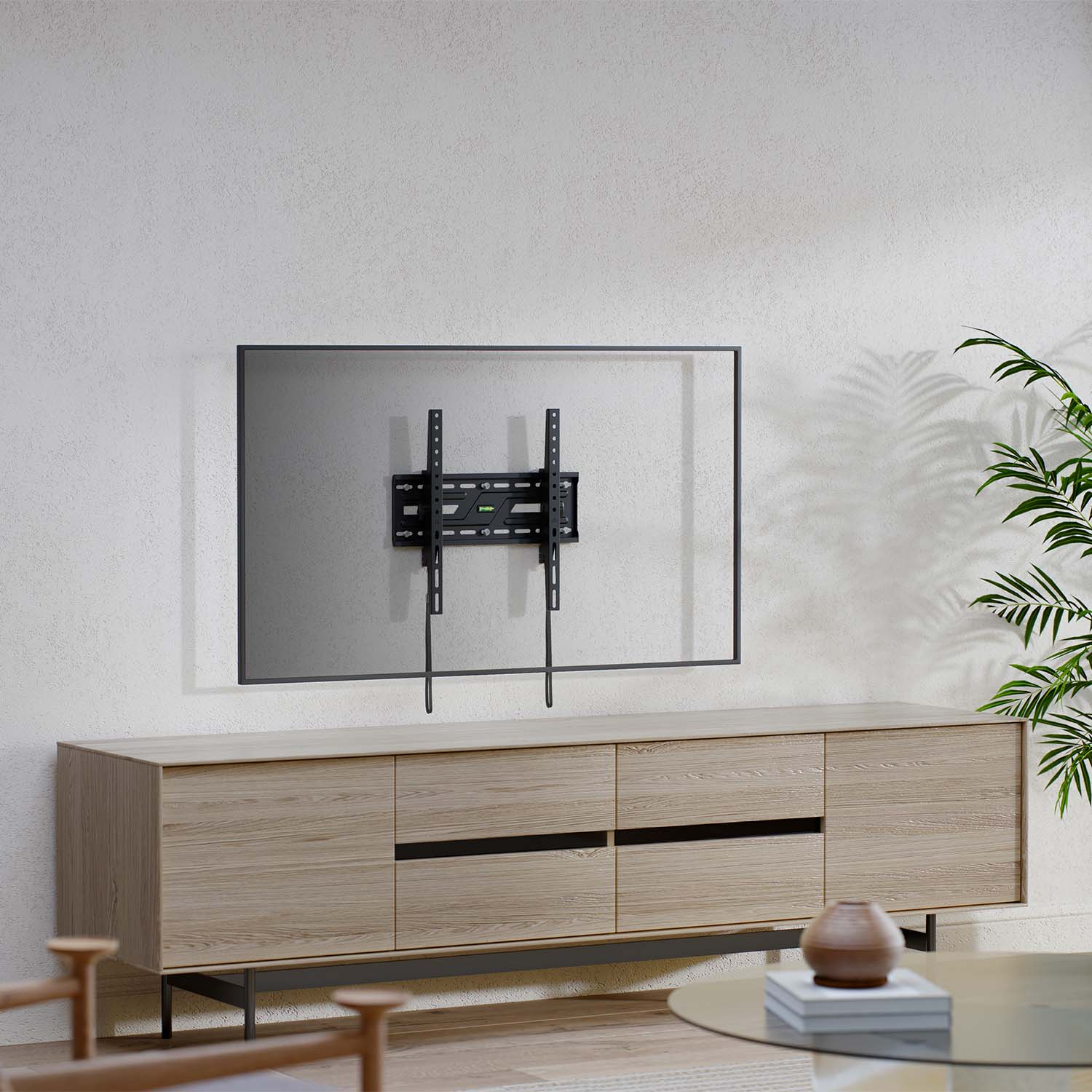 Un televisore è montato su un muro bianco sopra un mobile in legno. Una pianta è a destra. Un tavolino con libri è in primo piano.