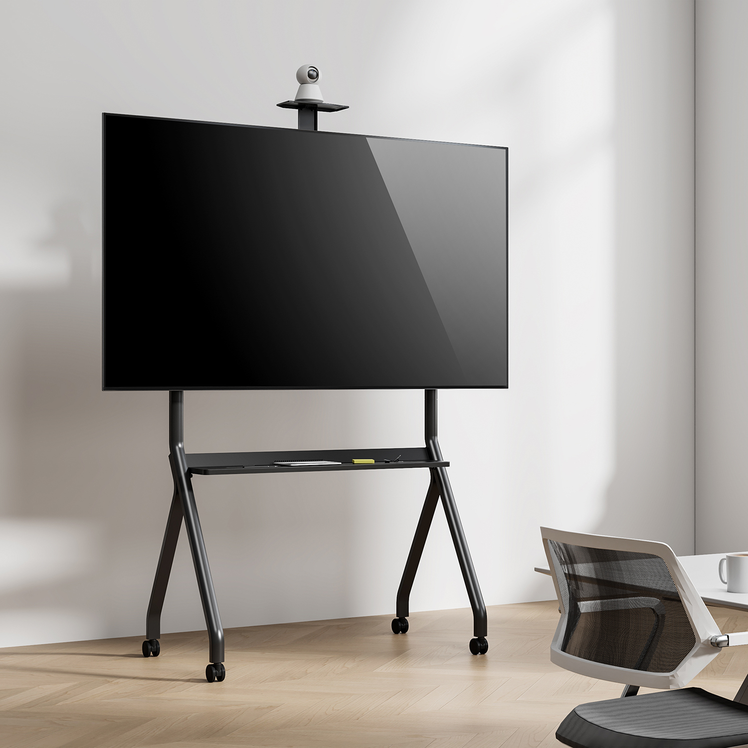 Una pantalla negra grande sobre un soporte negro con ruedas. Una cámara web en la parte superior. Una silla y mesa blancas a la derecha.