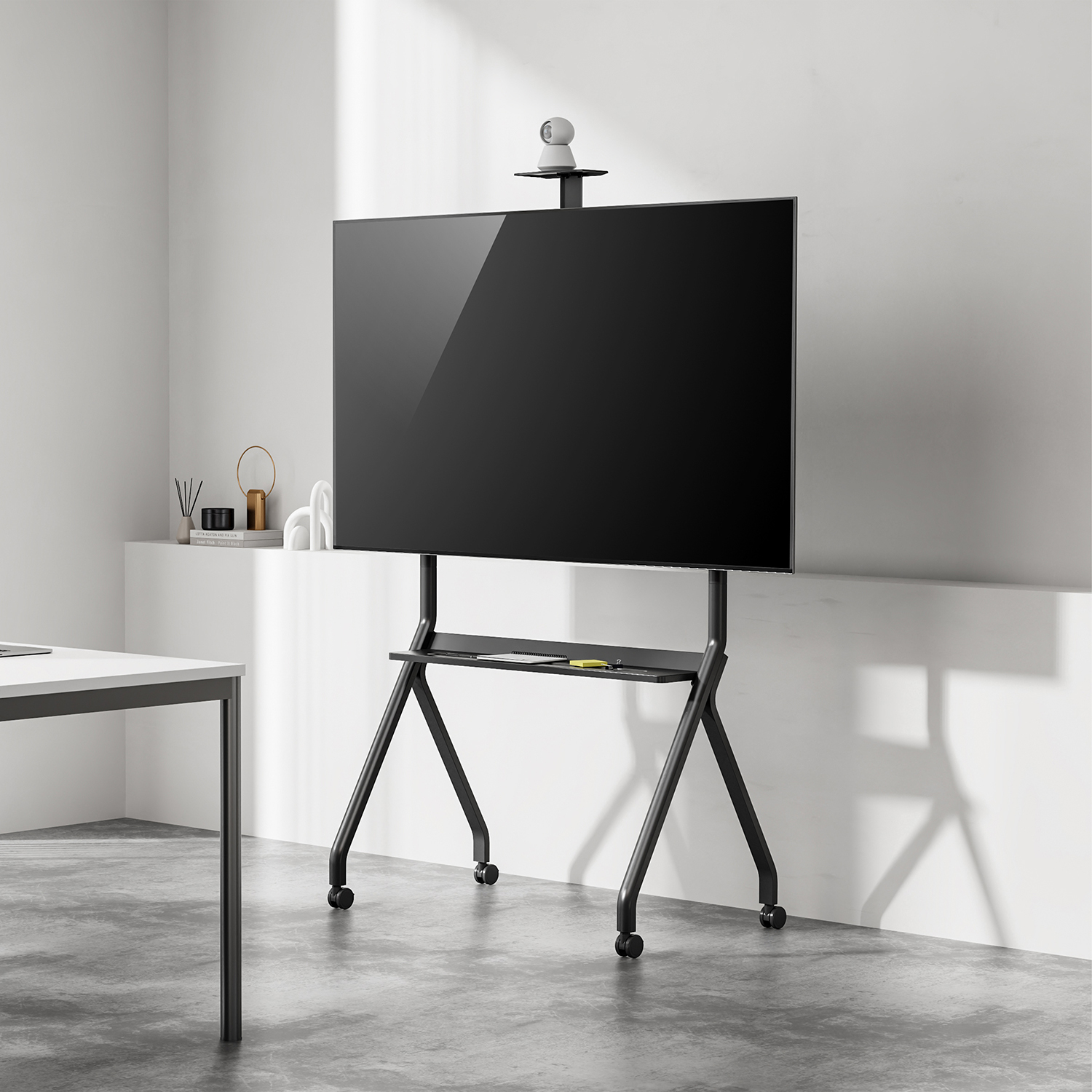 Un televisor negro sobre un soporte rodante. Una cámara encima. Un escritorio blanco y una pared blanca al fondo.