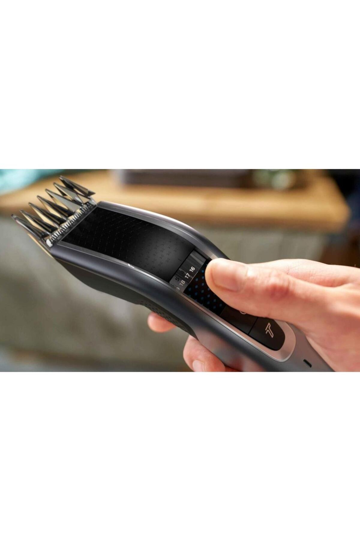 Dłoń trzymająca czarno-szarą maszynkę do włosów z widocznymi ustawieniami numerycznymi. Rozmyte drewniane tło.