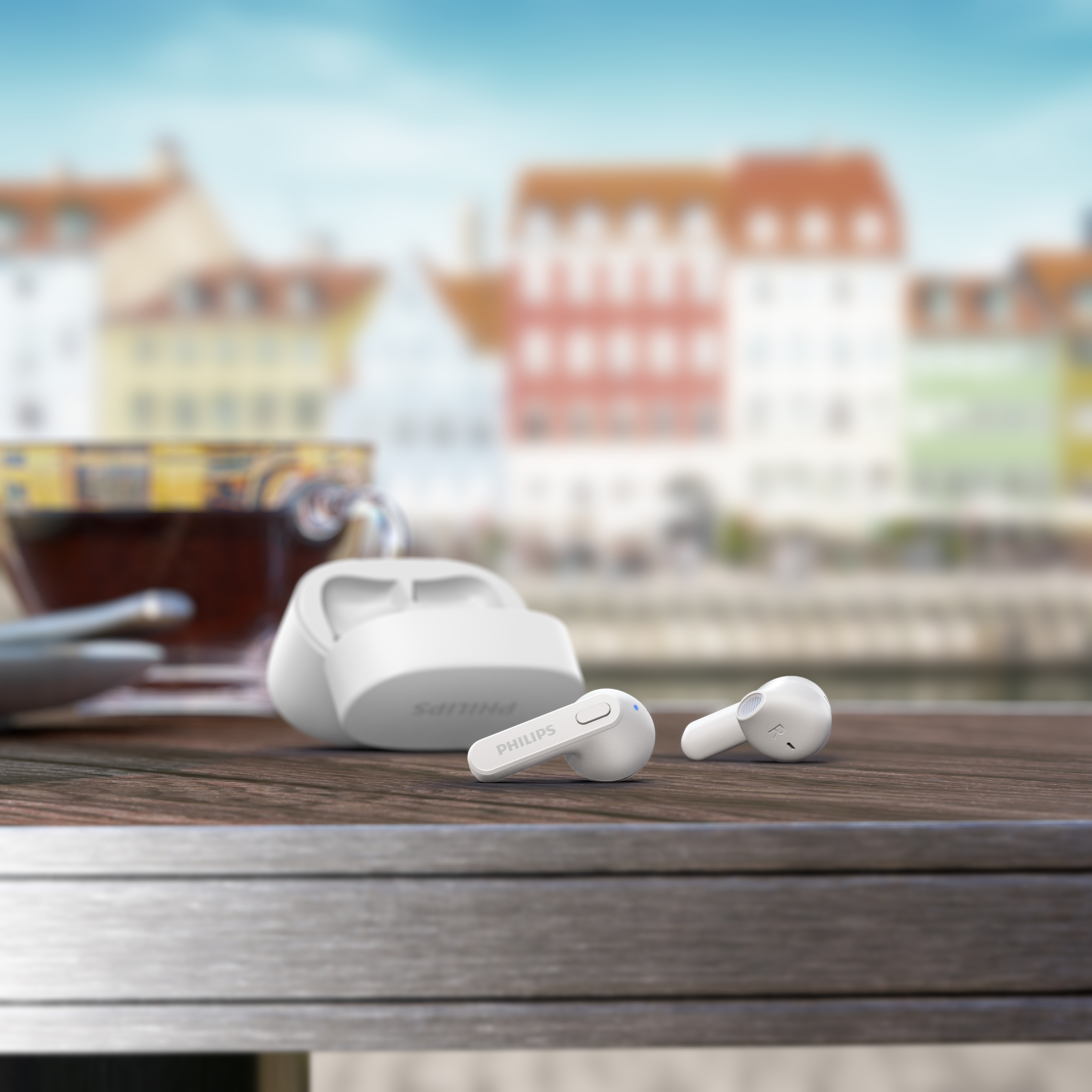 Auriculares y estuche de carga blancos de Philips sobre una mesa de madera, con un fondo urbano borroso y una taza de té.
