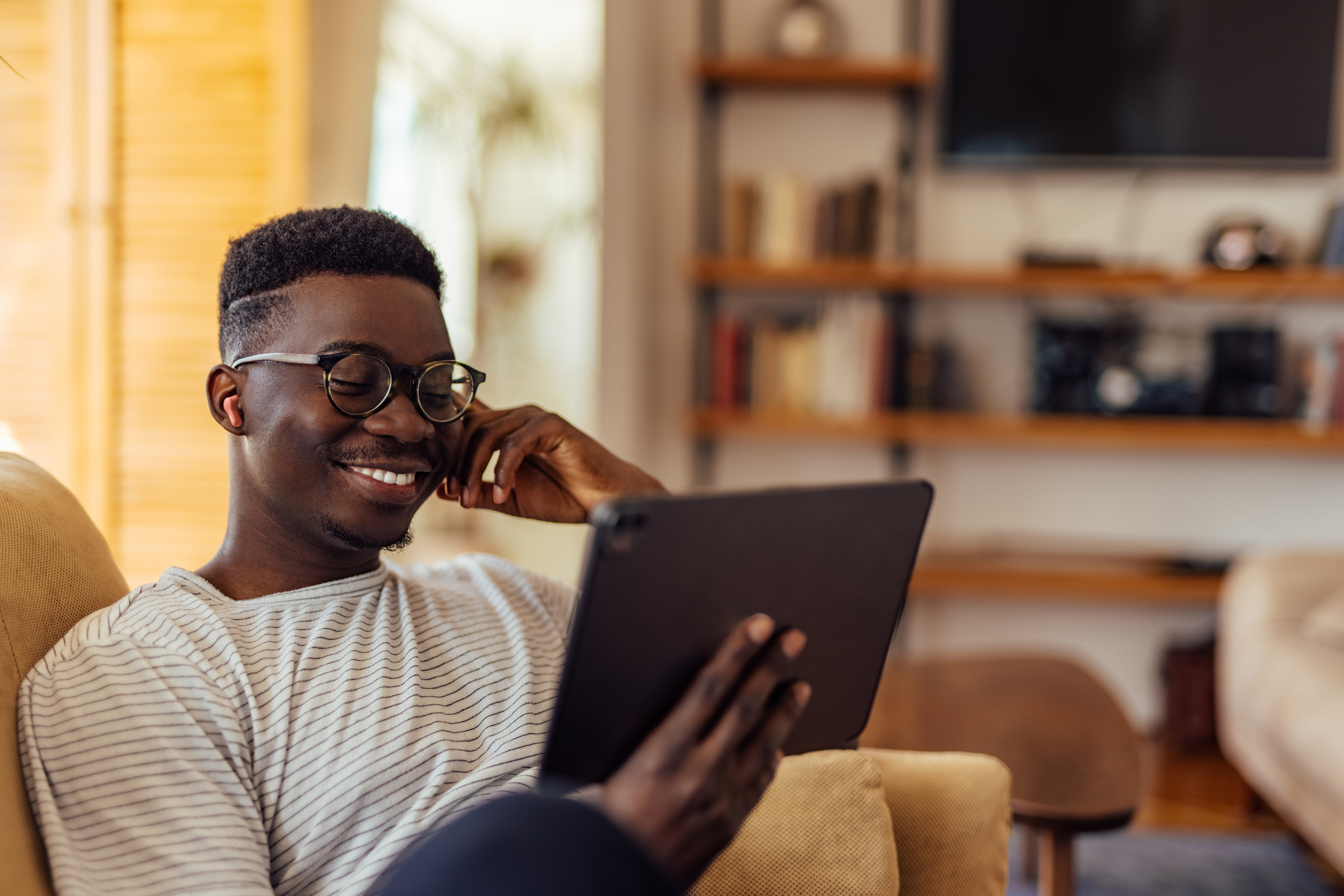 Un hombre negro sonriente con gafas sostiene una tableta. Está en una sala de estar con una estantería al fondo.