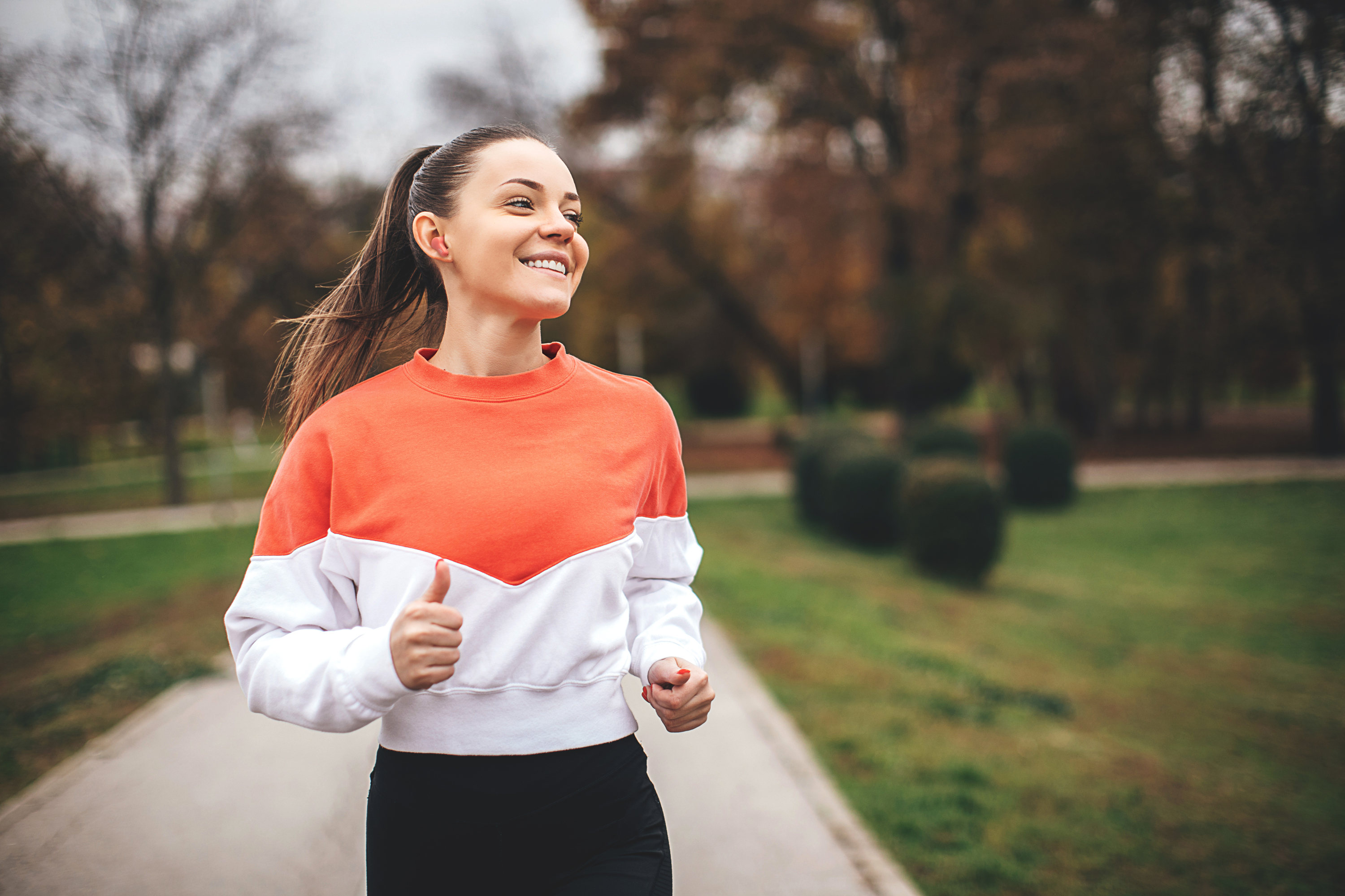 Una mujer sonriente corre por un camino, con una sudadera naranja y blanca. Árboles y vegetación en el fondo.
