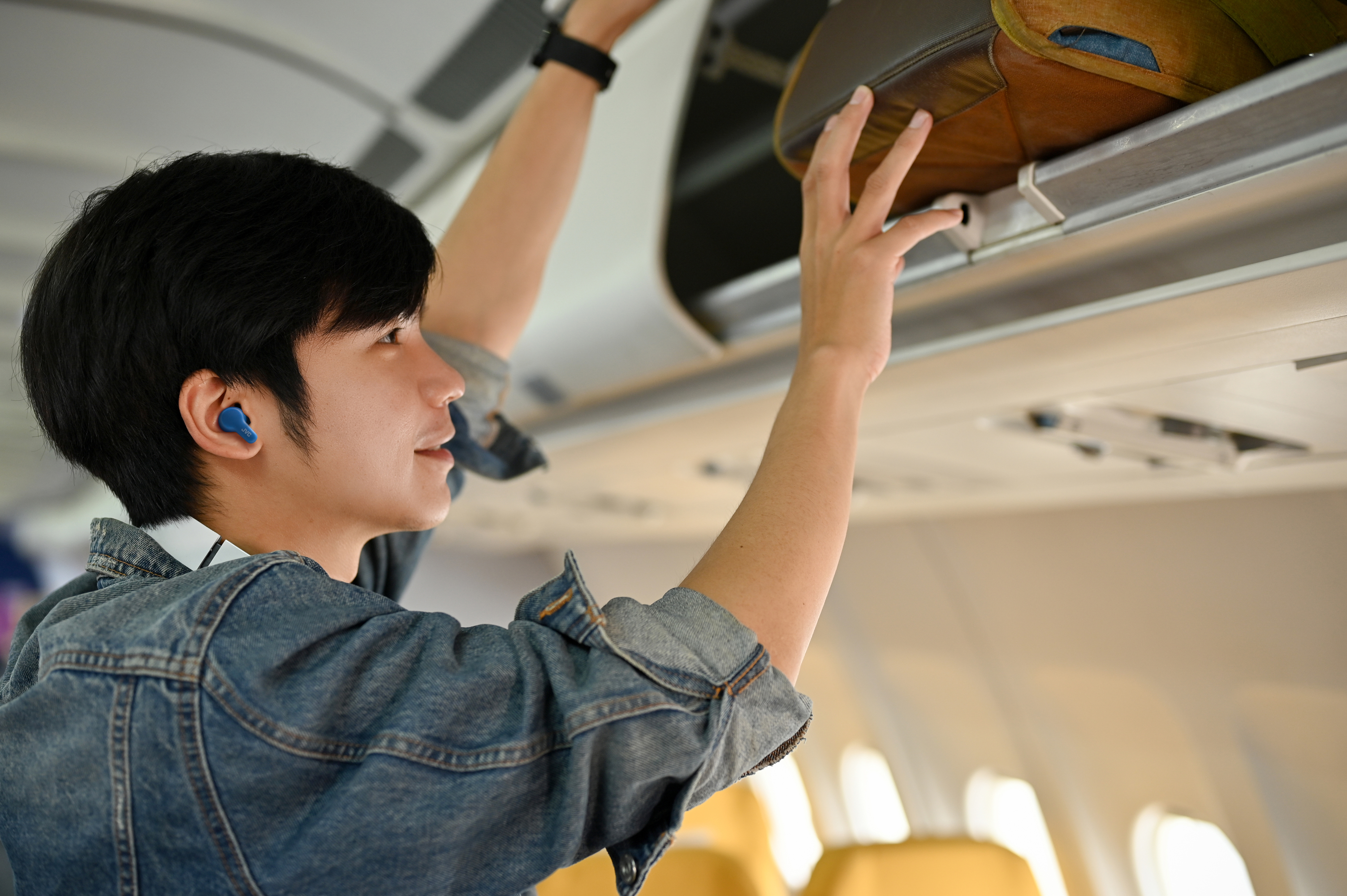 Un uomo con una giacca di jeans ripone i bagagli in un vano bagagli di un aereo. Ha auricolari blu.