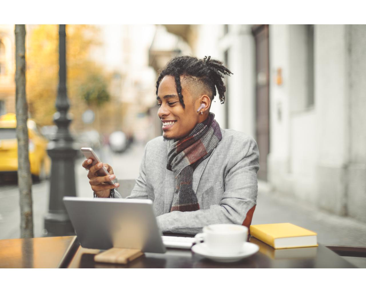 Mężczyzna uśmiecha się, używając telefonu, w szaliku i marynarce. Na stole laptop, książka i filiżanka kawy.