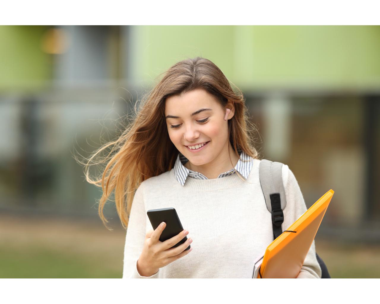 Młoda kobieta uśmiecha się, patrząc na telefon. Nosi plecak i pomarańczową teczkę. W tle budynek.