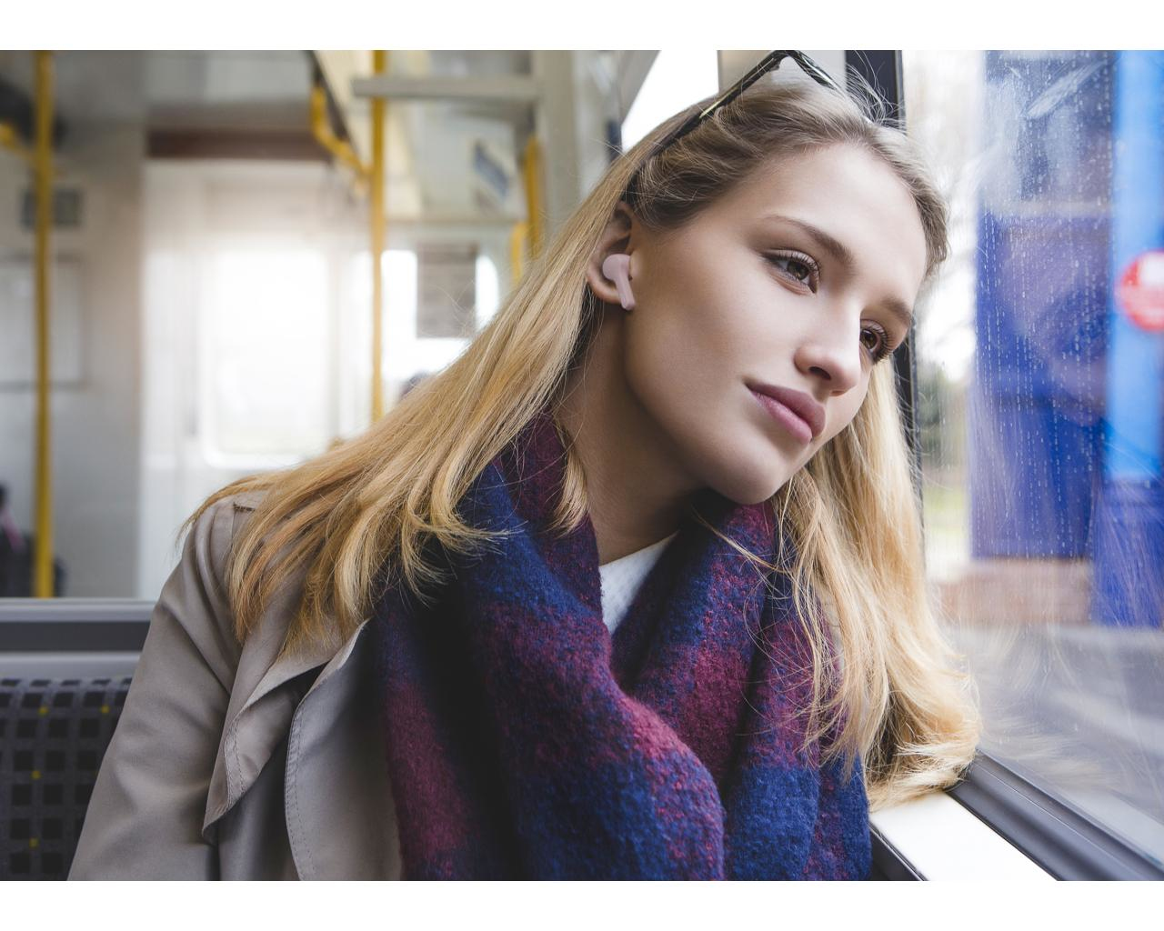 Blondynka w autobusie patrząca przez okno, w szaliku i słuchawce. Tło pokazuje siedzenia i jasne światło.