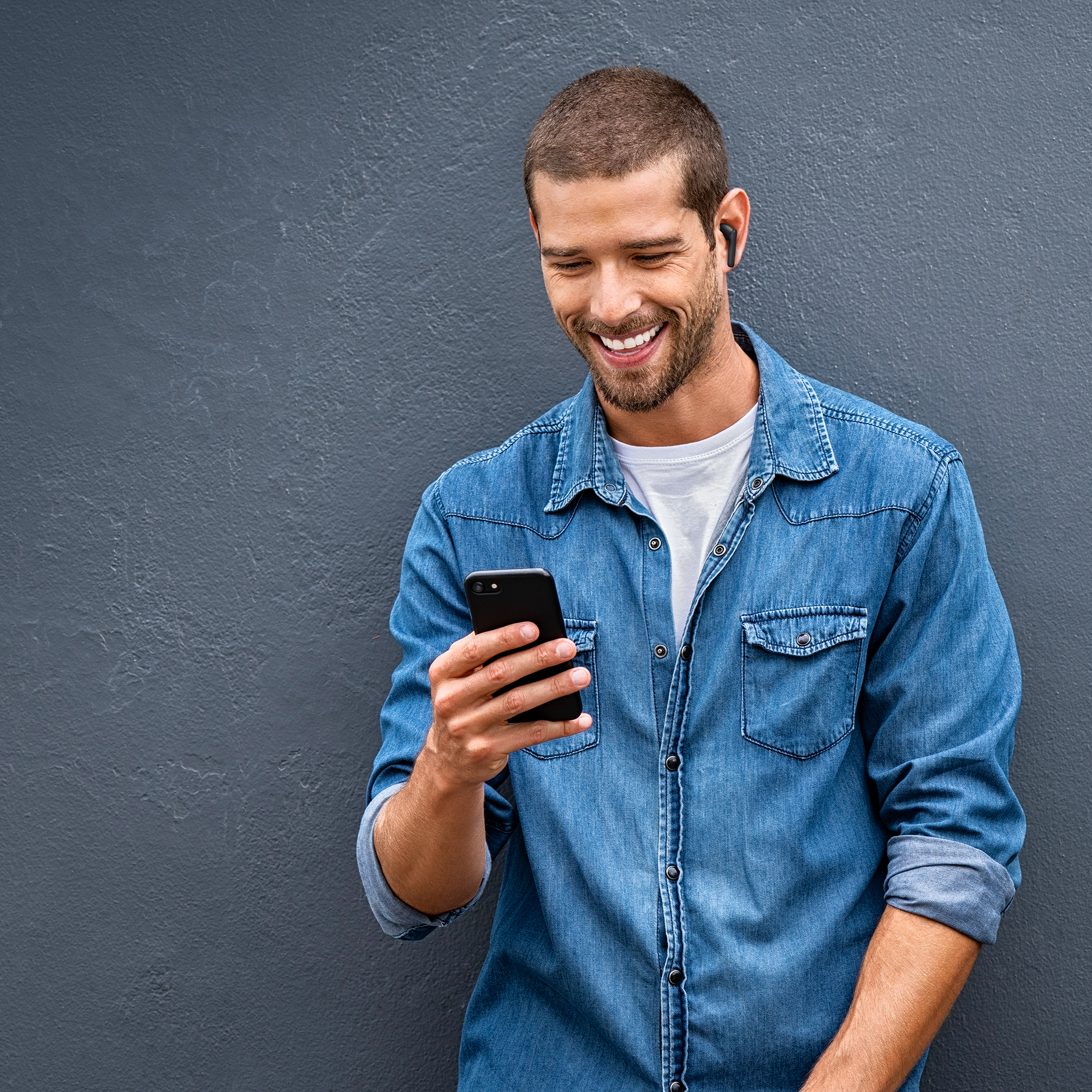 Ein lächelnder Mann in Jeanshemd schaut auf sein Handy vor einer grauen Wand. Er trägt Ohrhörer.