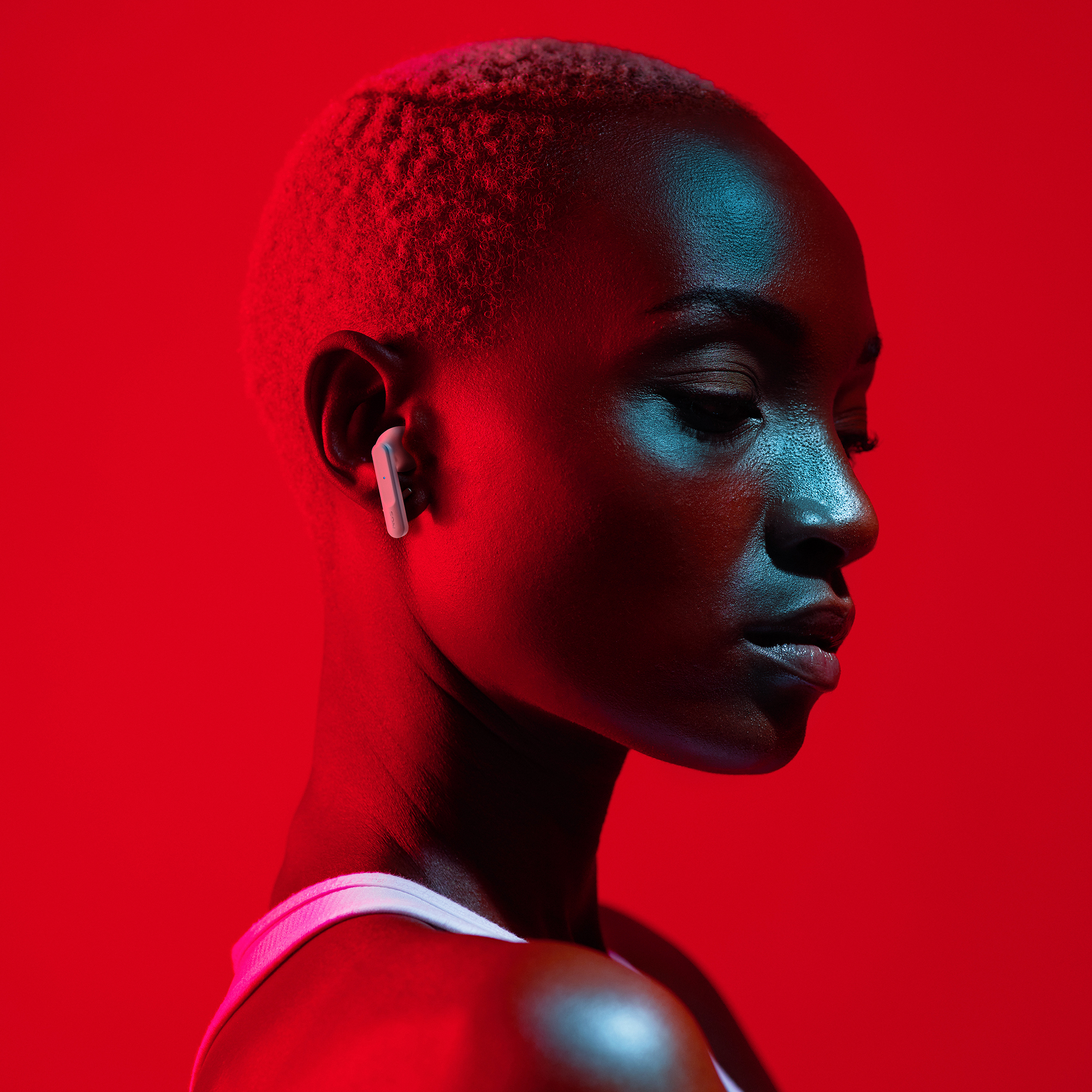 Una mujer negra con pelo corto usando un auricular, sobre un fondo rojo. Iluminación azul en la cara.